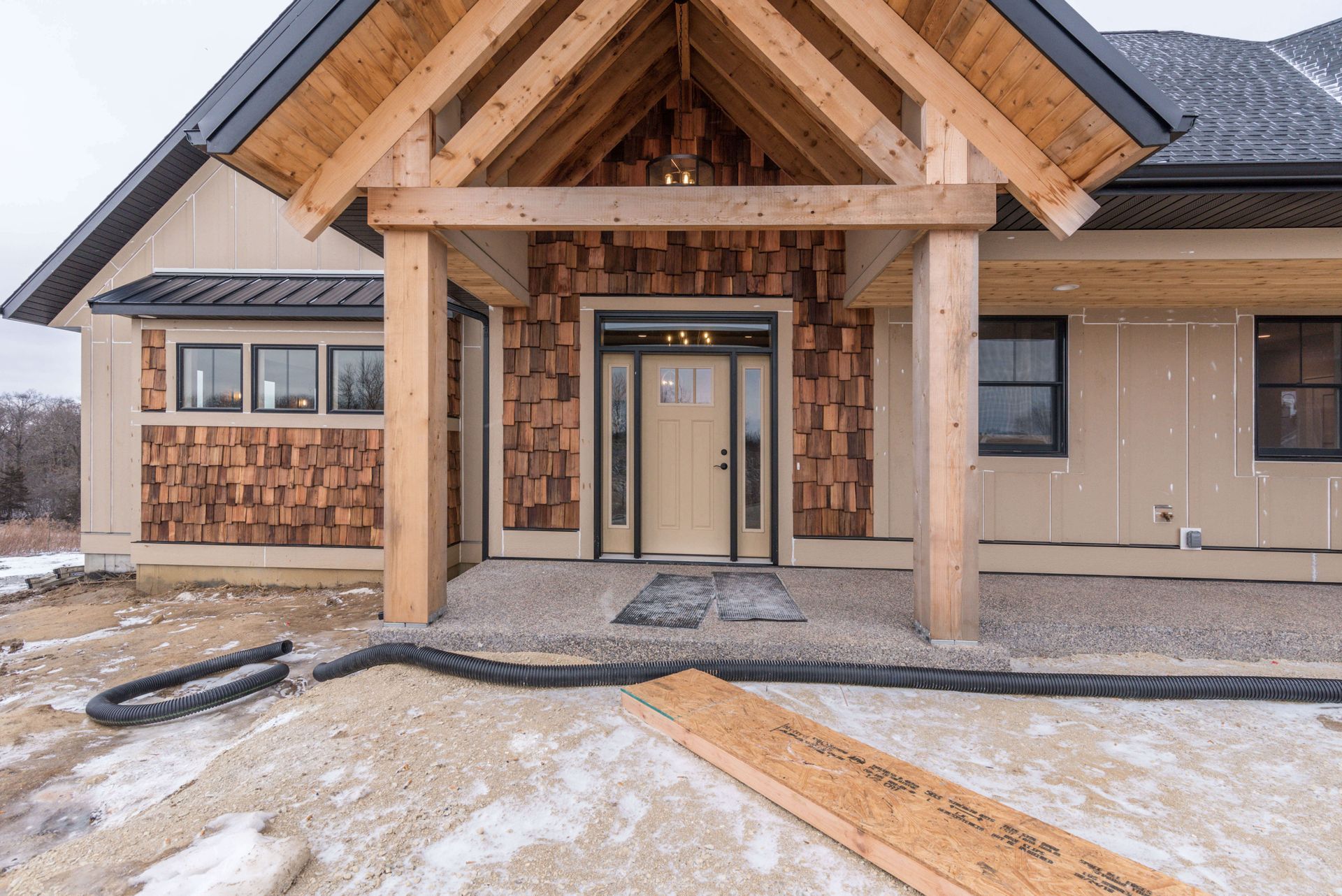 Entrance of a light-colored house with wooden posts and a cedar shake accent wall; snow and gravel in the foreground.