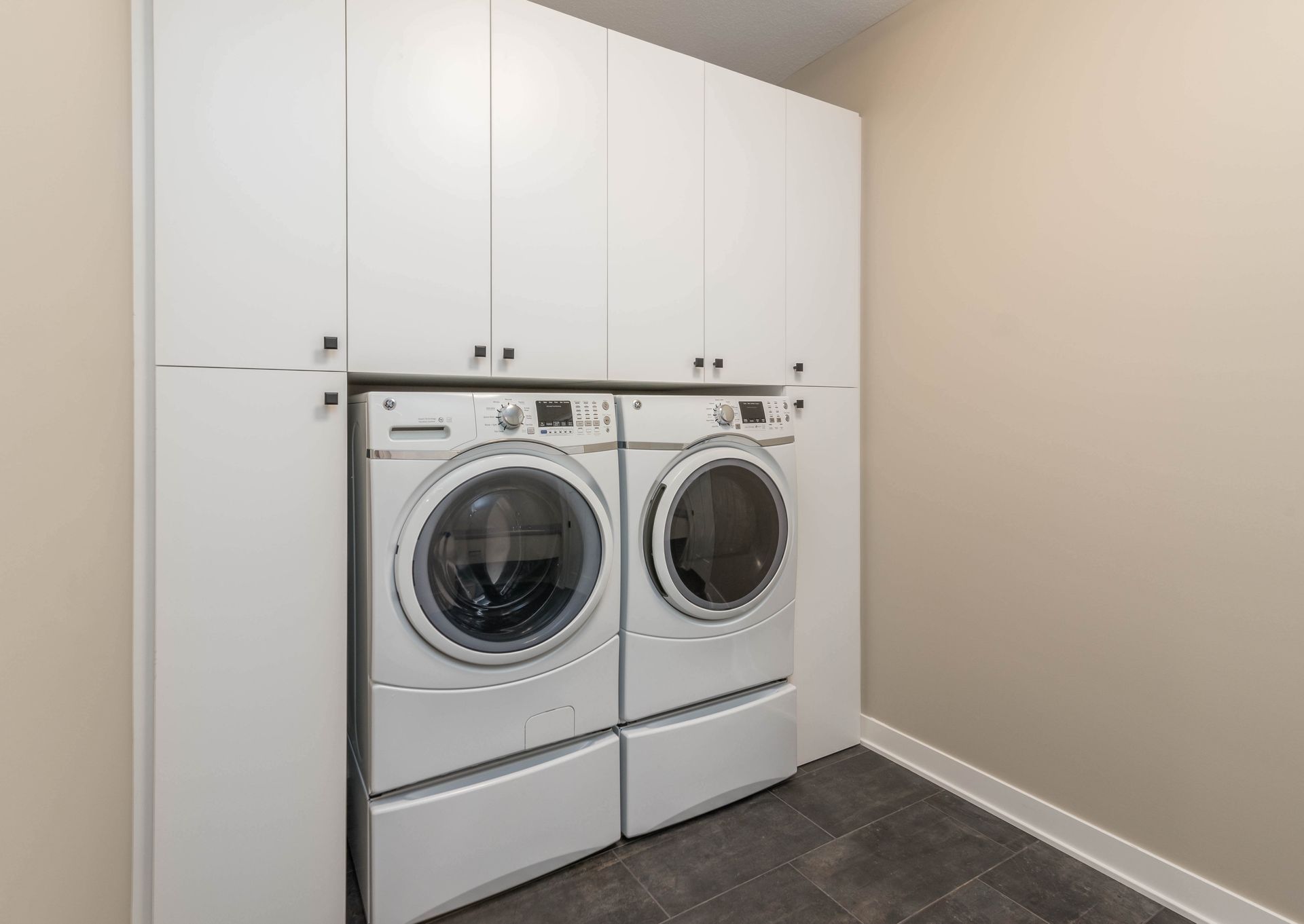 White laundry machines beneath white cabinets in a beige-walled laundry room with dark tile flooring.