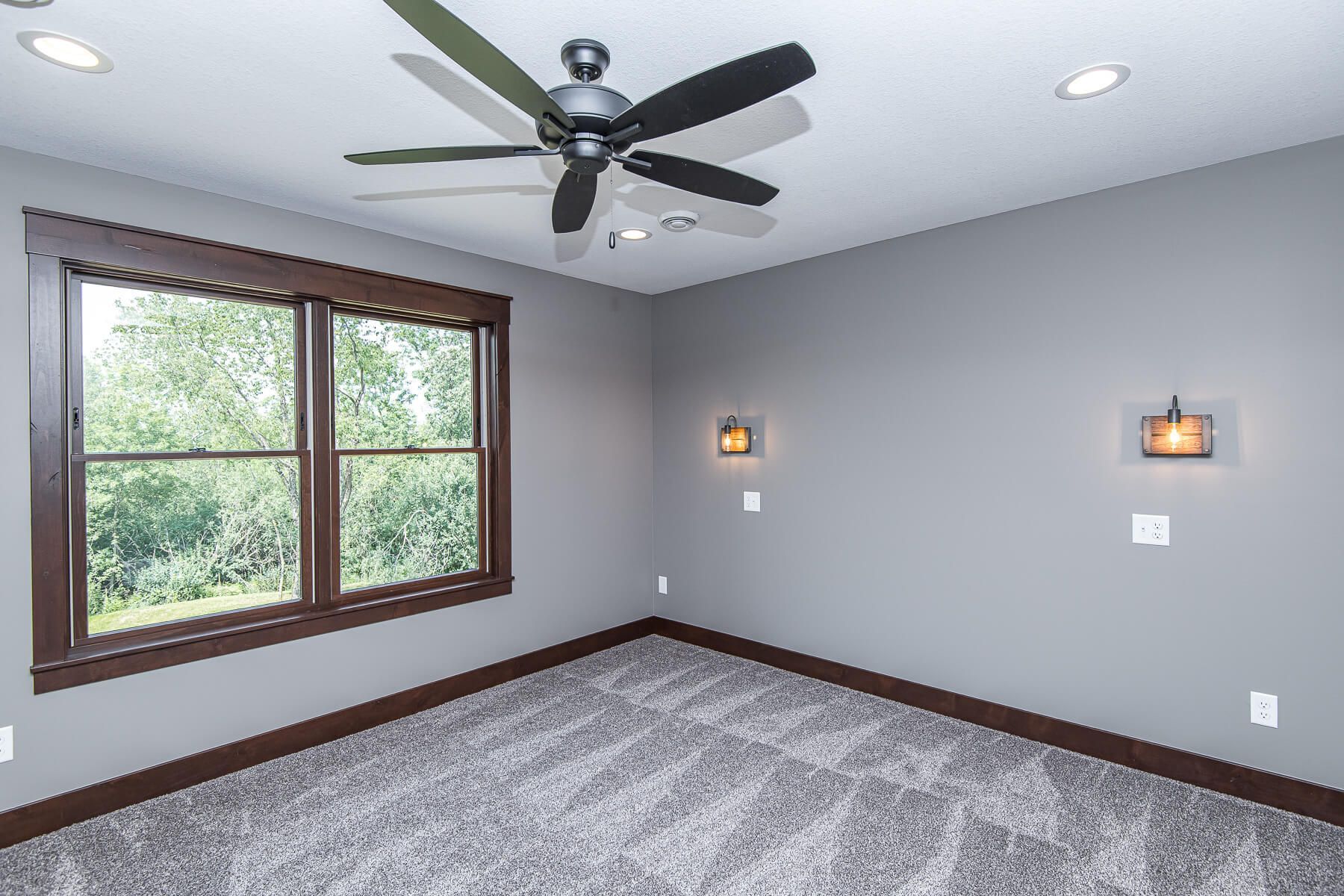Empty bedroom with gray walls, brown trim, and a window. Ceiling fan and sconces.