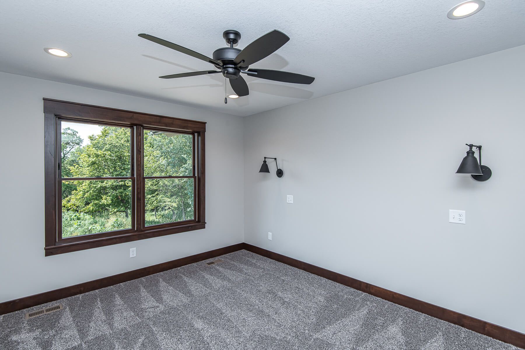 Bedroom with gray carpet, walls, dark wood trim, ceiling fan, sconces, and window overlooking trees.