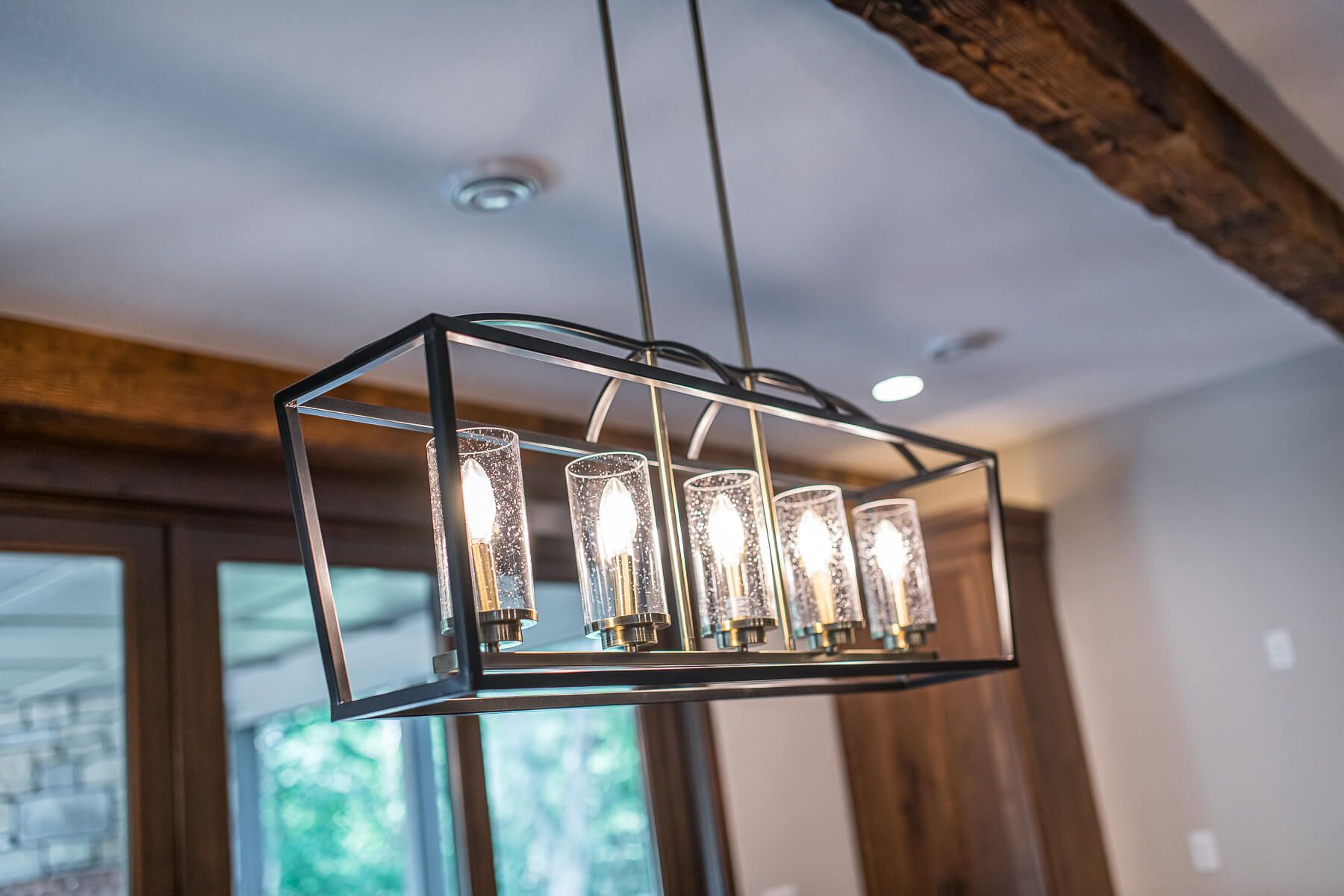 Modern chandelier with five glass shades, hanging in a room with wood beams and a window.
