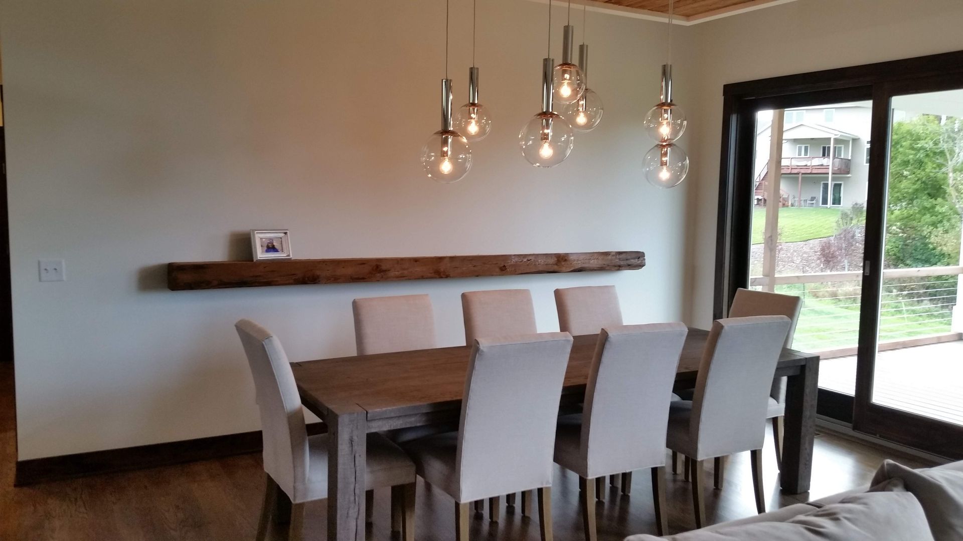 Dining room with wooden table, eight white chairs, a wooden shelf, and pendant lights. A sliding glass door overlooks a yard.