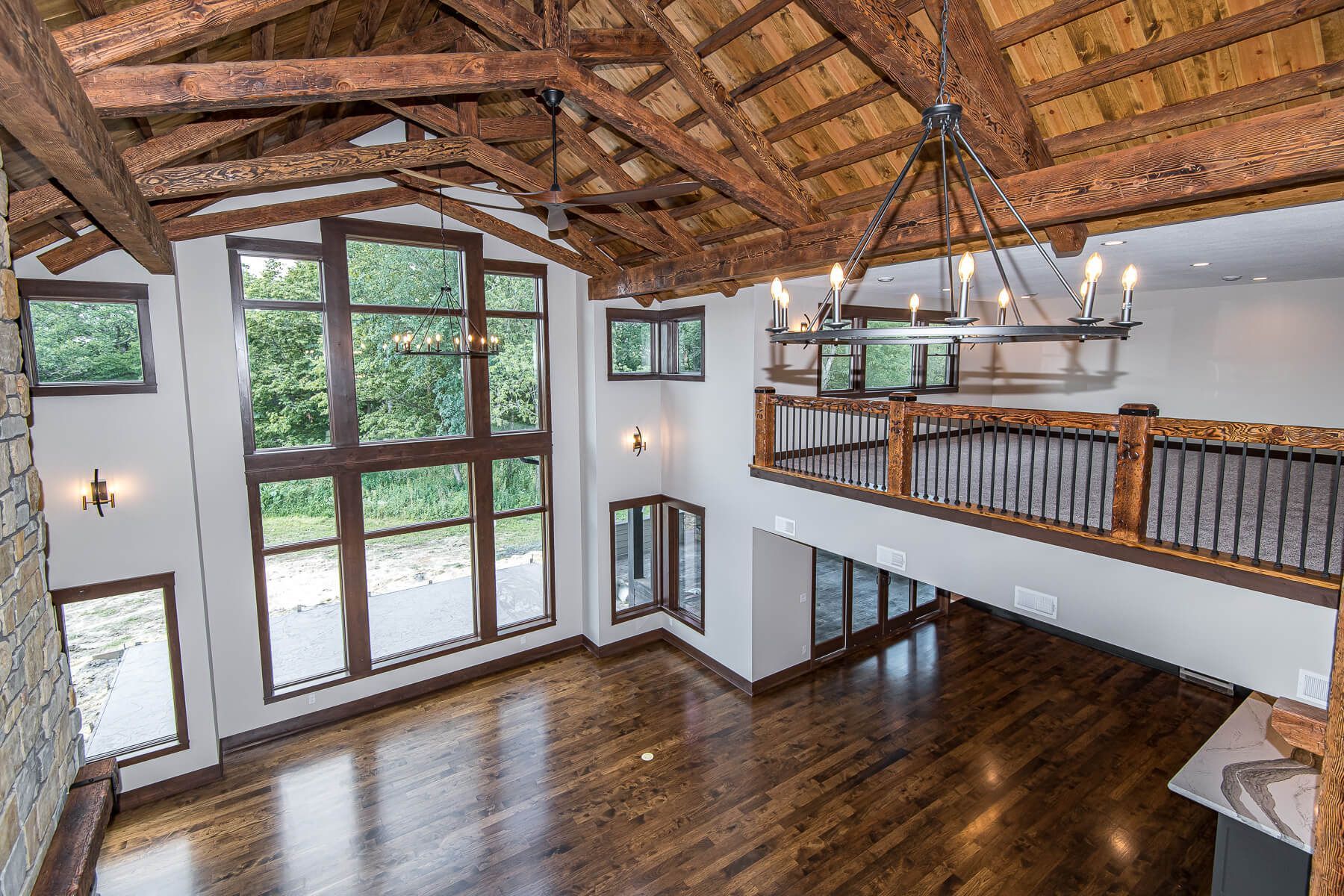High-ceilinged room with wood beams, large windows, wooden floor, balcony, and chandelier.