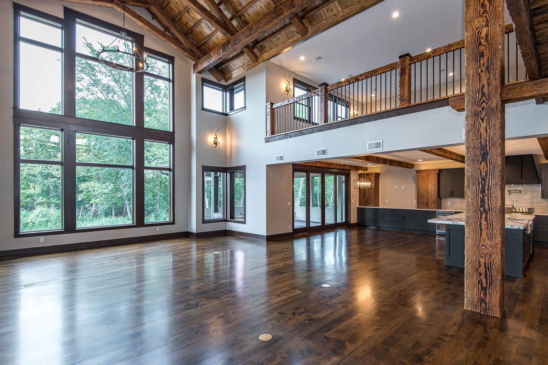 Spacious interior with exposed wooden beams, large windows, and hardwood floors. A loft overlooks the open space.