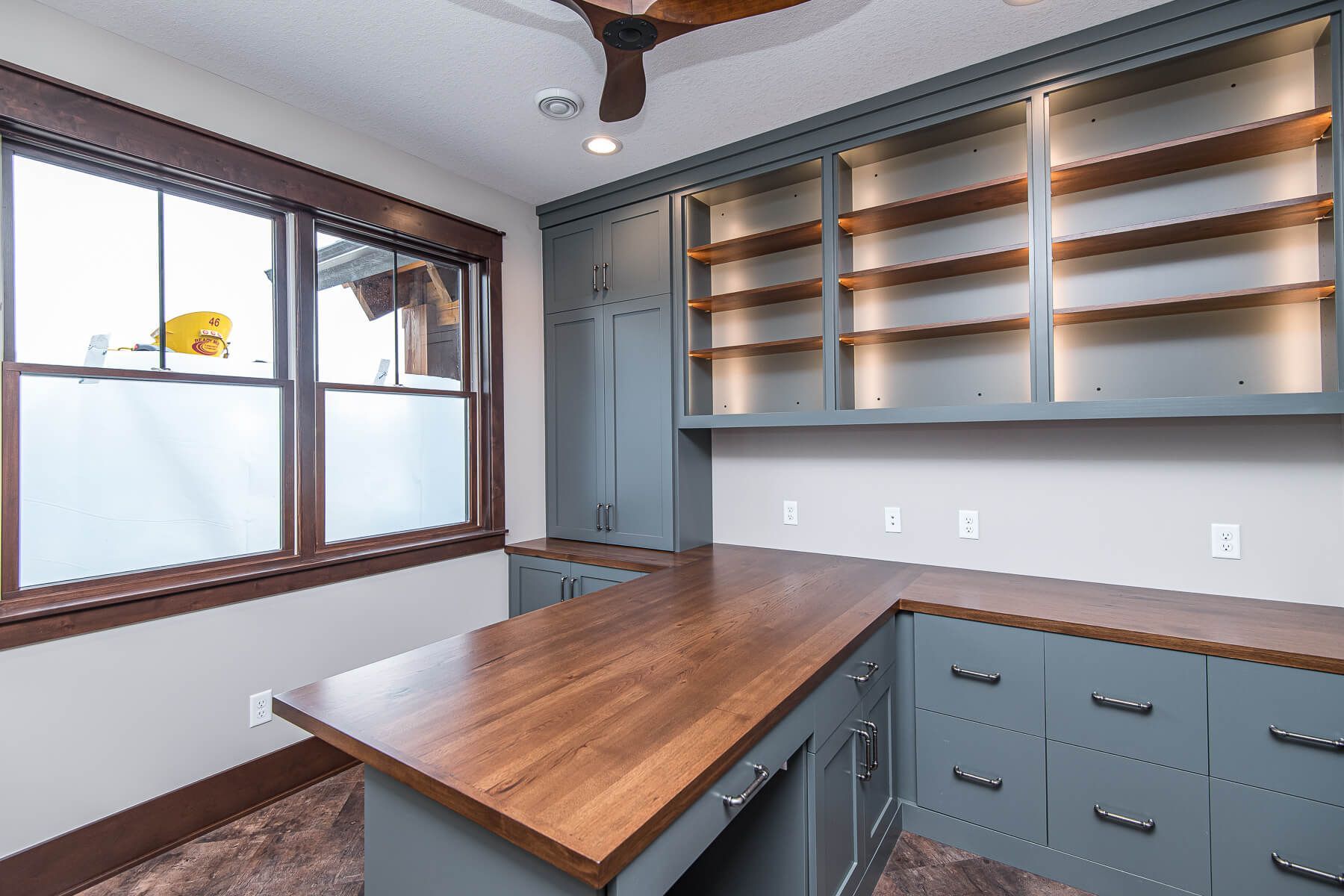 Office with blue cabinets, wood countertop, and window with brown trim. Empty shelves.