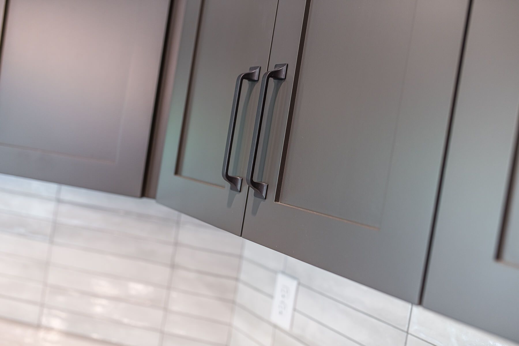 Gray kitchen cabinets with black handles above white tiled backsplash.