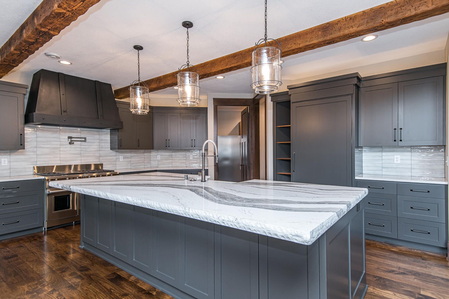 Modern gray kitchen with island, pendant lights, wooden beams, stainless steel appliances, and white countertops.