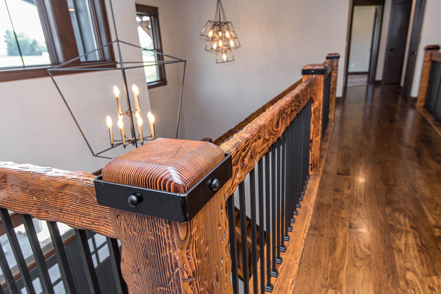 Wooden banister with black metal spindles and hardwood floor in a home hallway, lit by multiple chandeliers.