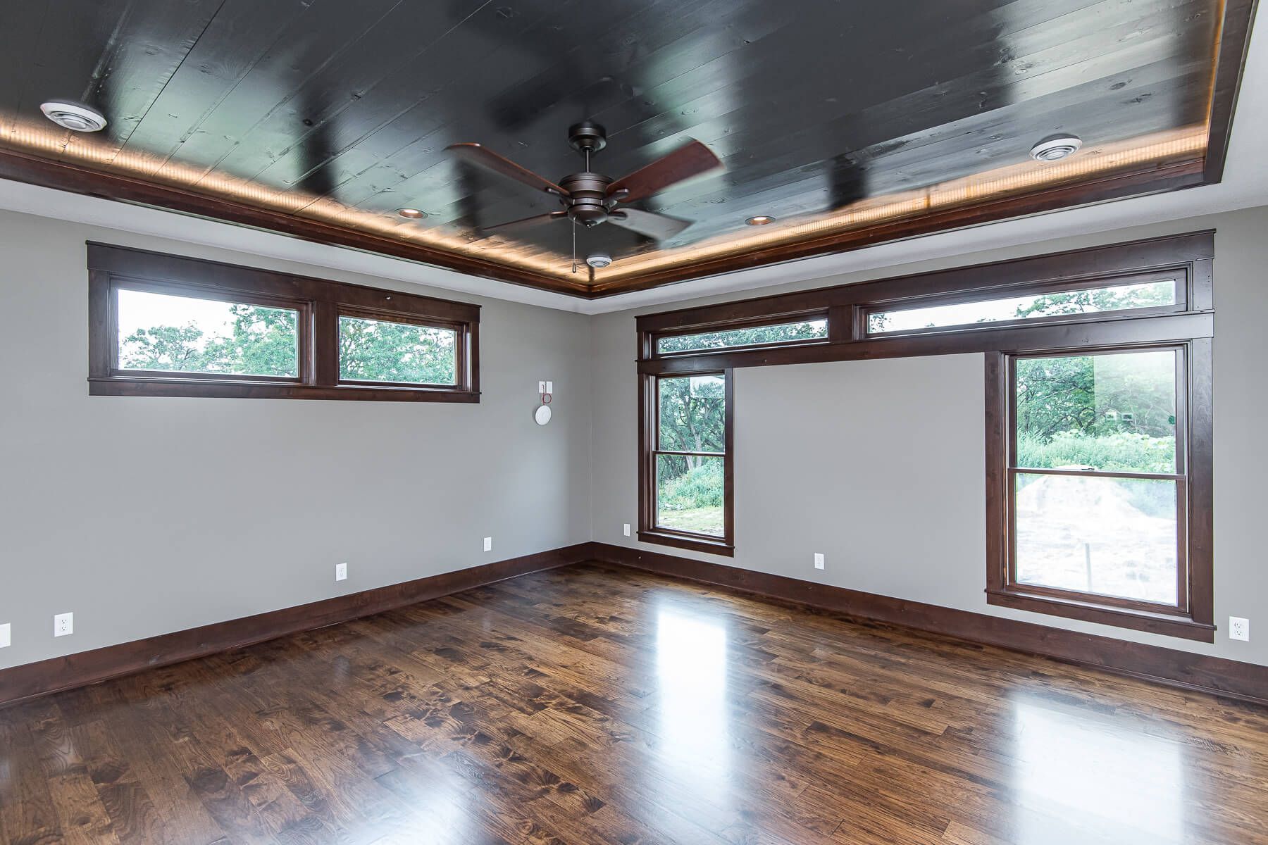 Empty room with dark wood ceiling, trim, and floor. Gray walls and large windows with outdoor views.