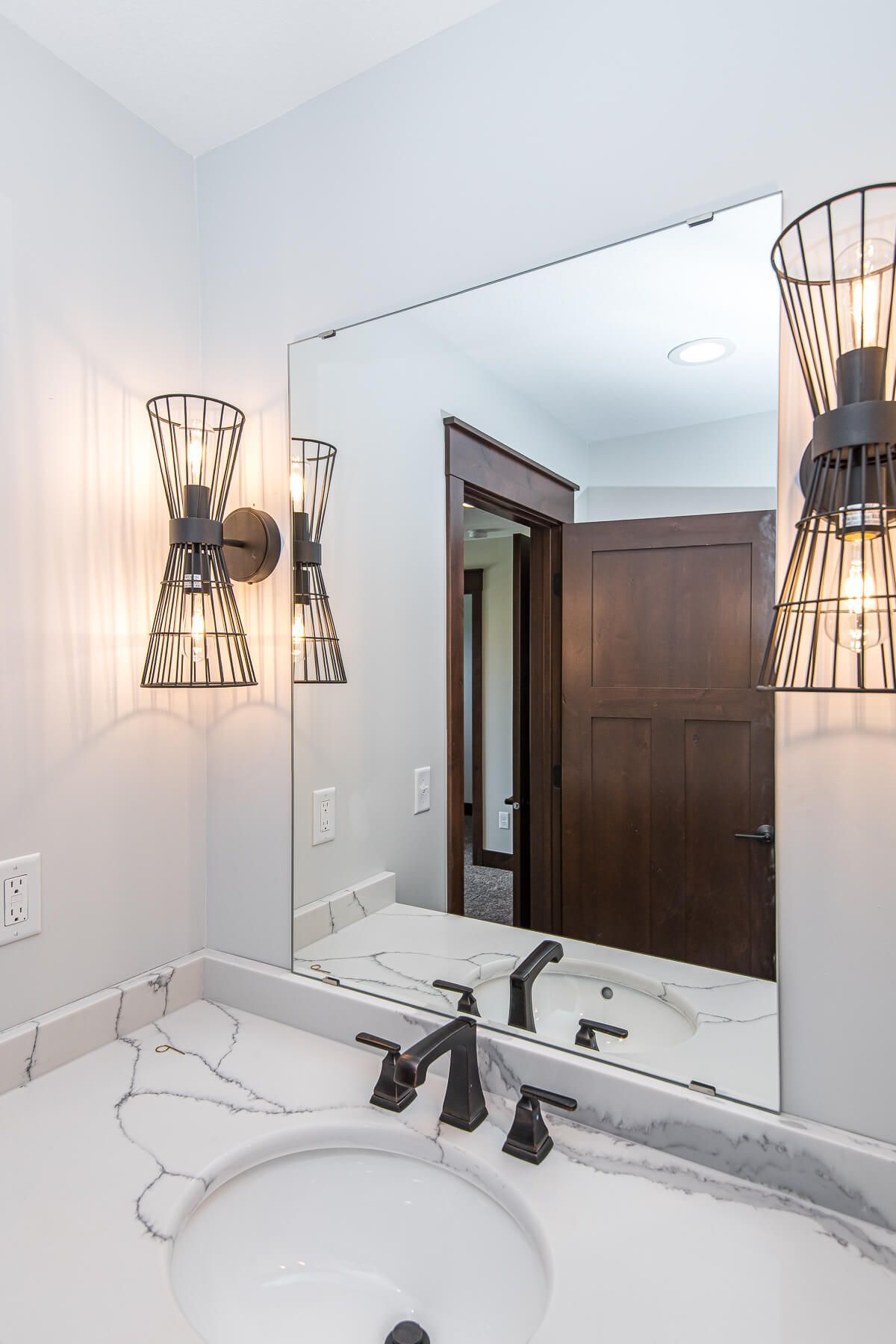 Bathroom vanity with a white countertop, large mirror, and dark sconces. Dark wood door visible.