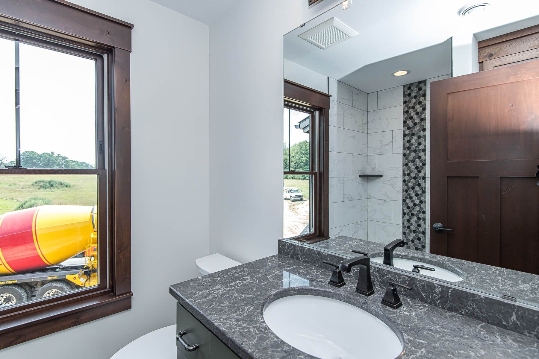 Bathroom with dark wood trim, gray countertop, and a large mirror. A concrete mixer is seen outside the window.