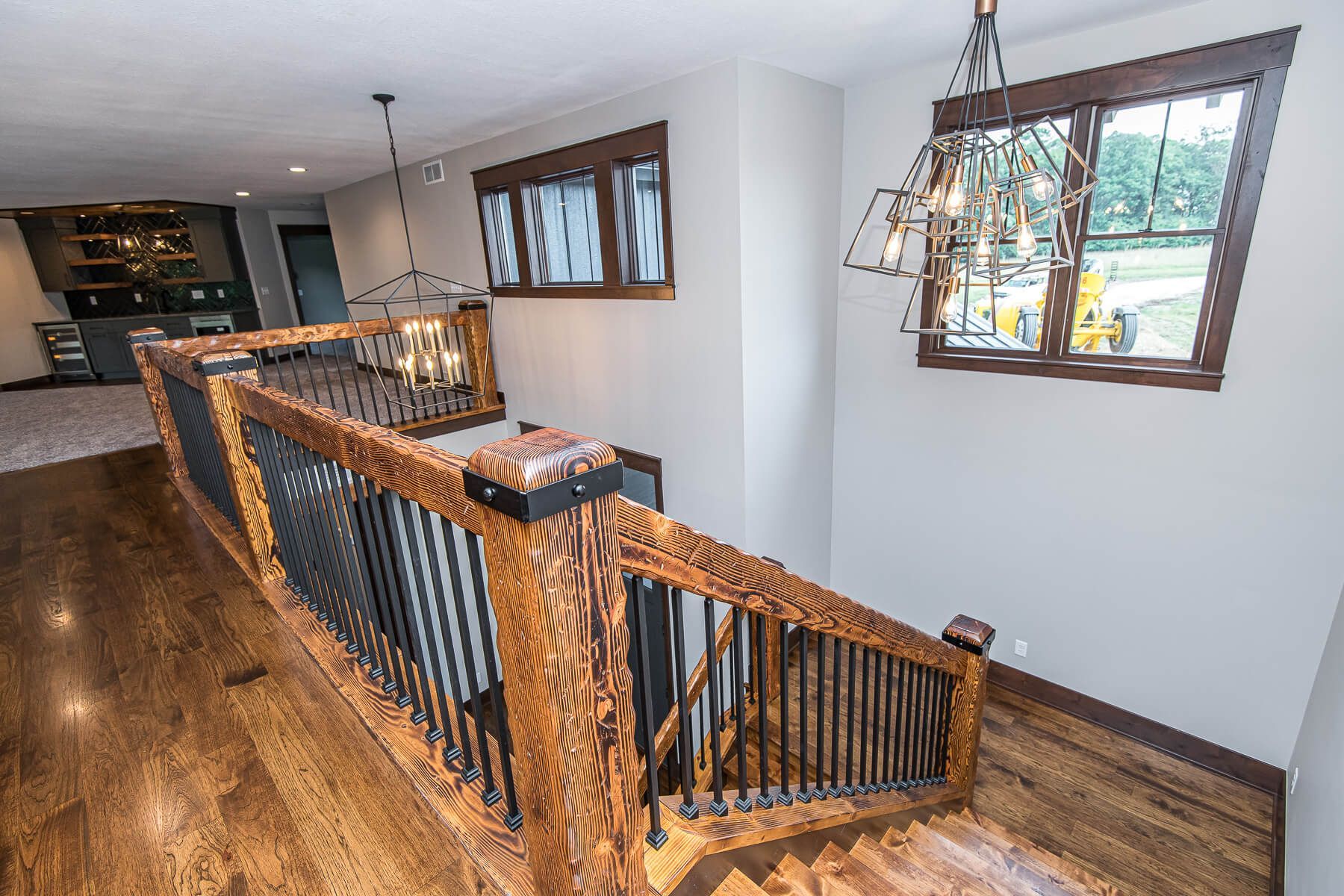 Wooden staircase with black railings, two chandeliers, and a view of a bar and window.