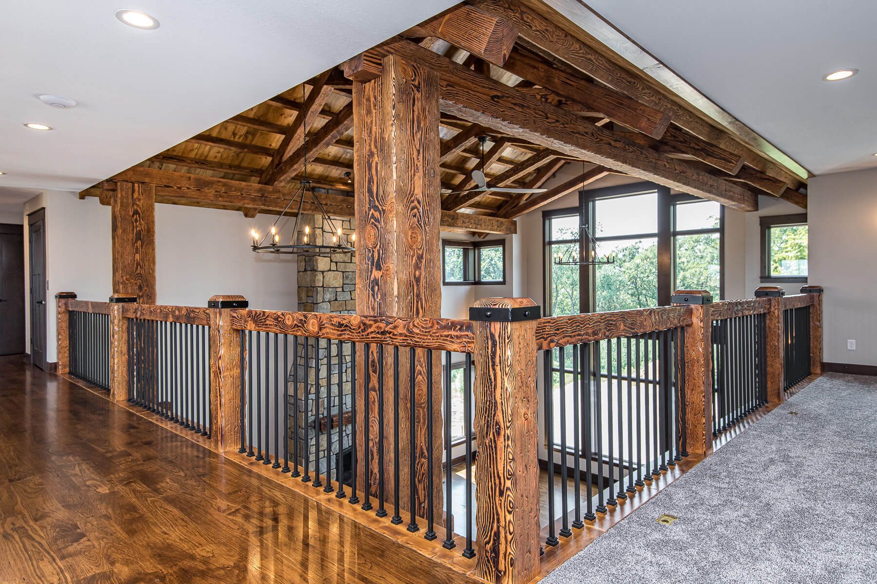 Wooden balcony with dark iron railing, overhead beams, and a view into a room with a chandelier.