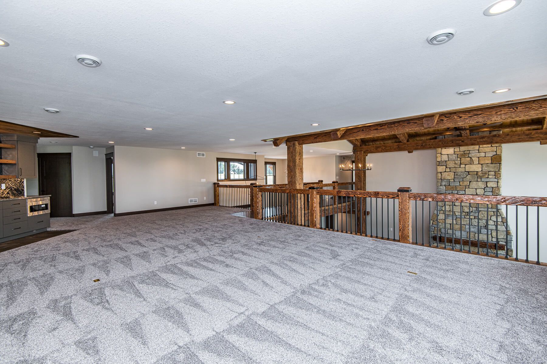 Spacious room with gray carpet, wood beams, and a stone accent wall. Overhead lighting and railing present.