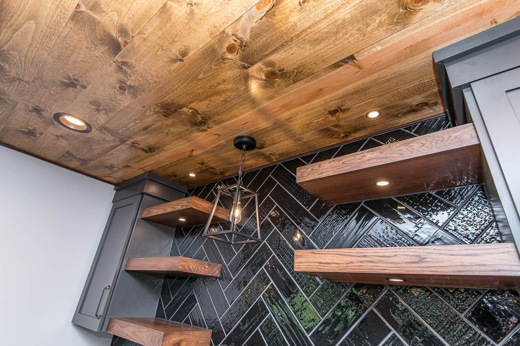 Wooden ceiling and shelves against a black herringbone-patterned tile wall; pendant light hangs.