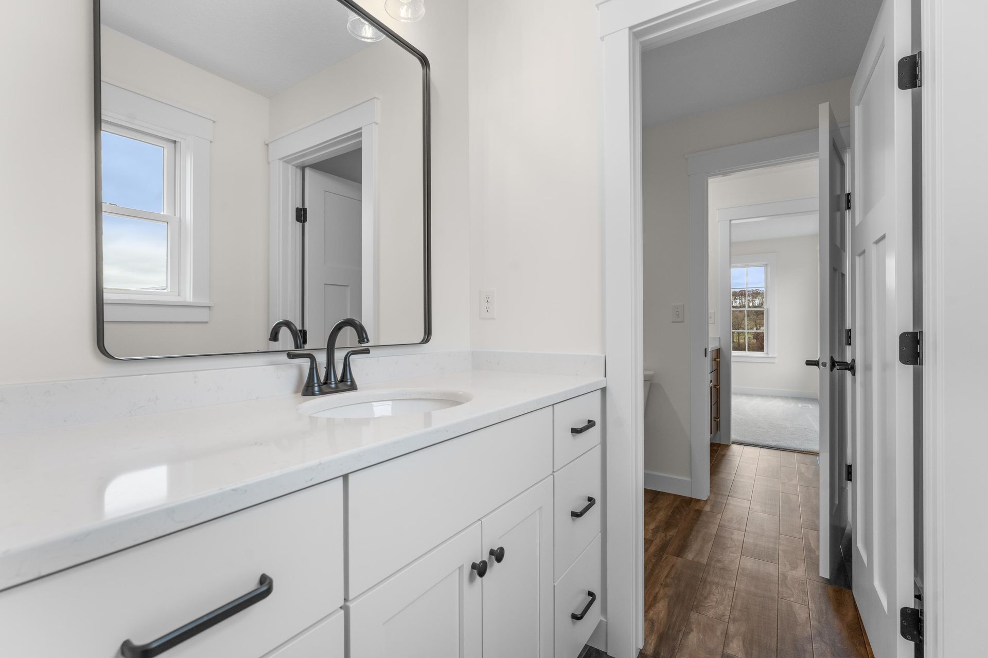 White bathroom with dark fixtures, mirror, and open doorway to hallway with wood flooring.