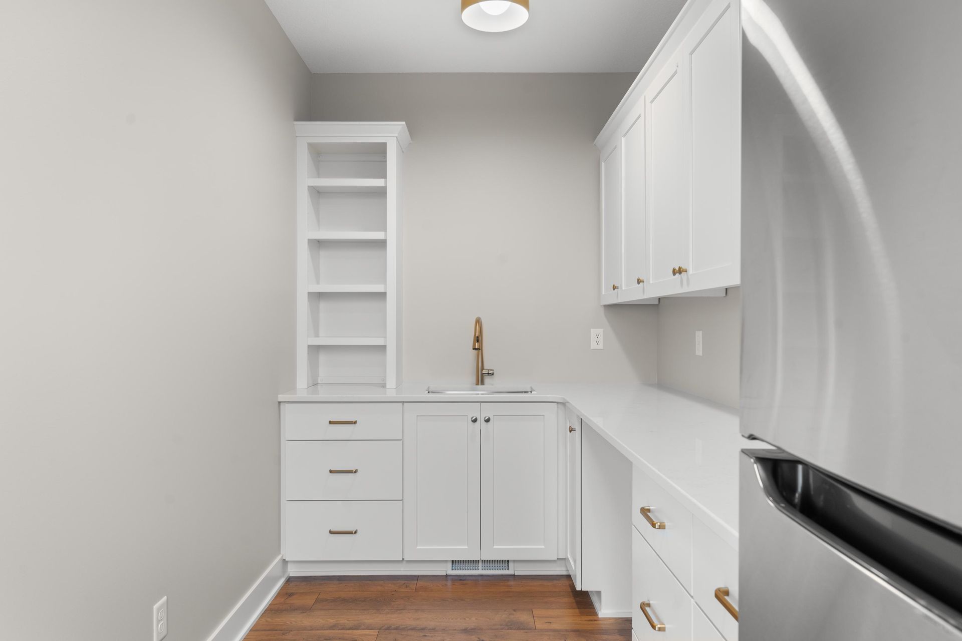 White cabinetry with sink and open shelving in a small kitchen area; stainless steel refrigerator visible.