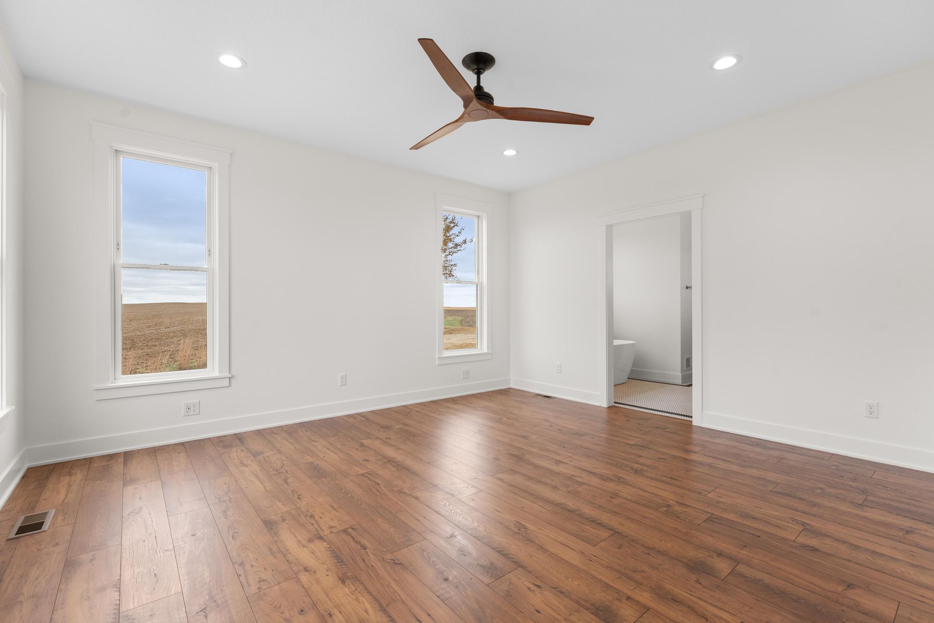 Empty, bright bedroom with hardwood floors, windows, and a ceiling fan. An open doorway leads to a bathroom.