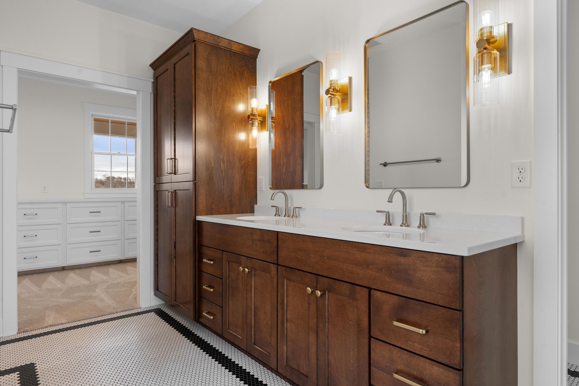 Bathroom with dark brown vanity, white countertop, mirrors, and sconces; doorway to a room with white cabinets.