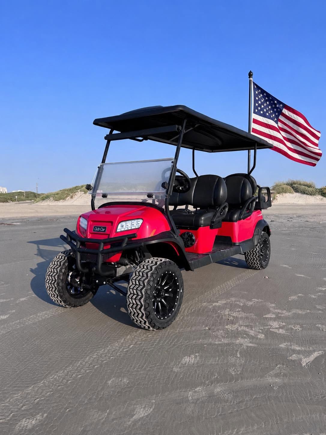 Red off-road utility vehicle with black roof and American flag parked on a sandy road near dunes