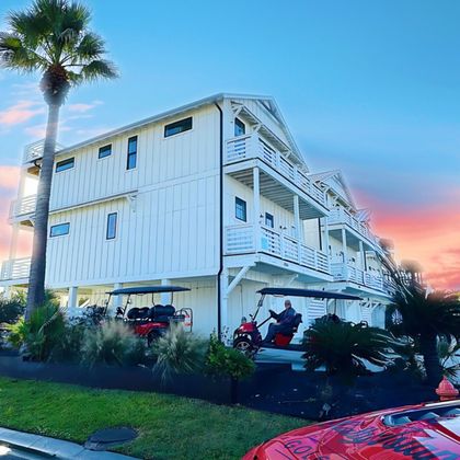 White beach house with palm trees at sunset, red car in front, and two motorbikes parked below.