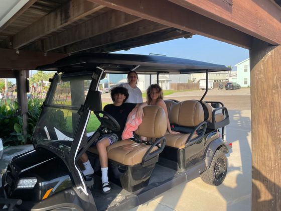 Three people sitting in a tan golf cart under a shaded carport.