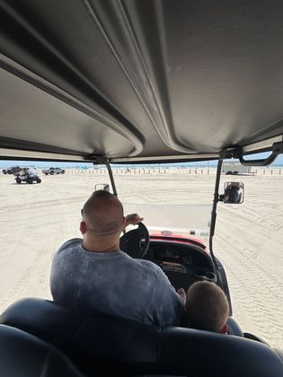 Two people ride in an open off-road vehicle across a sandy desert, viewed from the back seat.