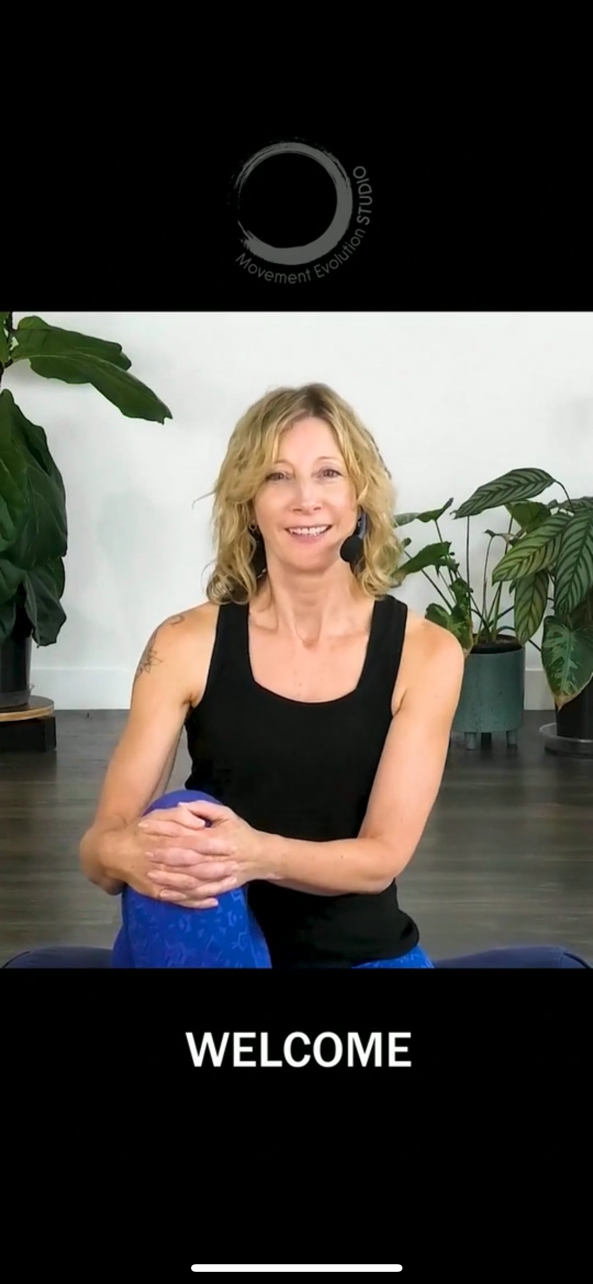 A woman in a black tank top is sitting on a yoga mat. movement evolution studio mobile app screen shot
