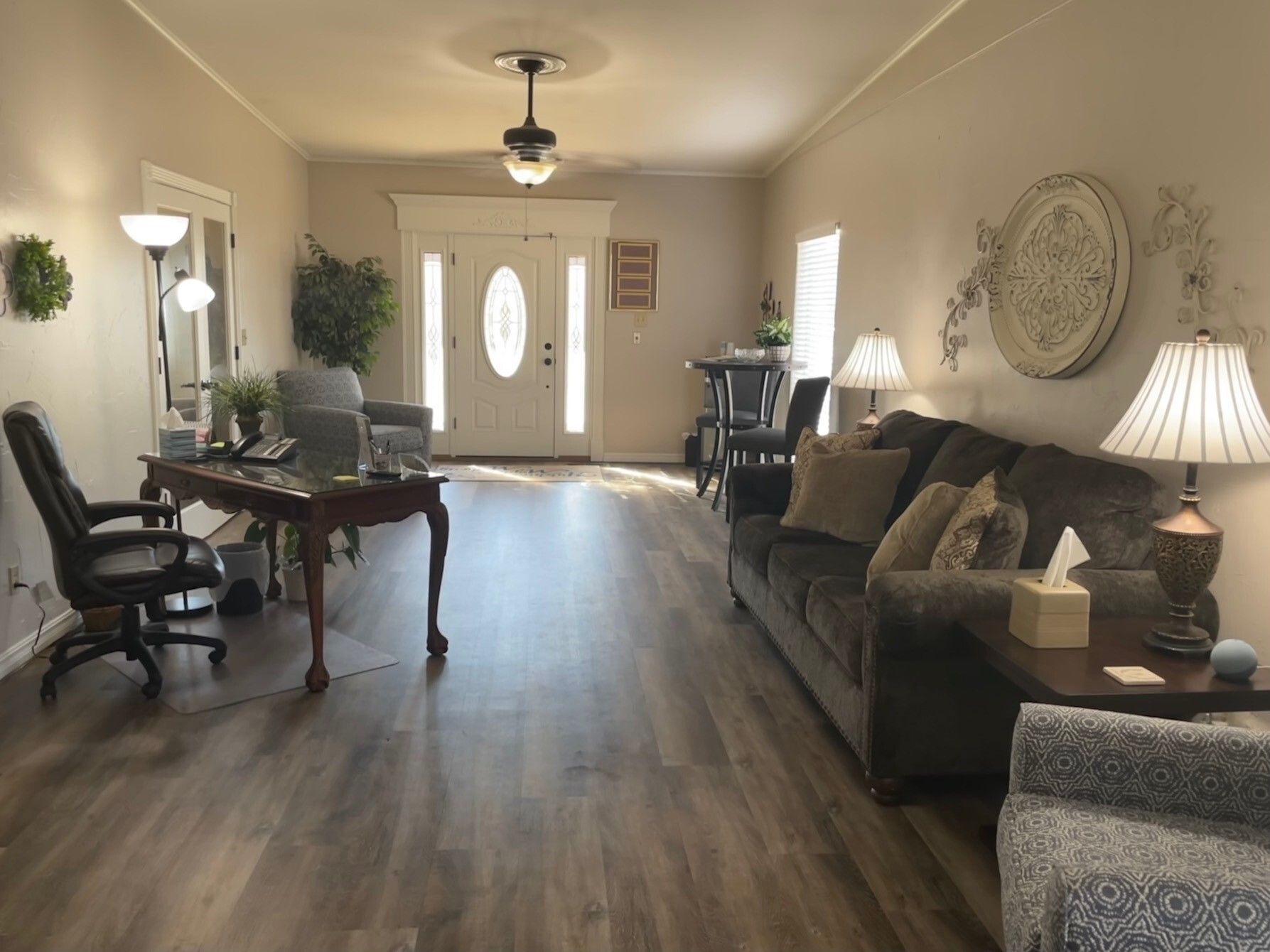 Bright living room with gray sofa, desk, wood floors, front door, and lamps in a tidy open space