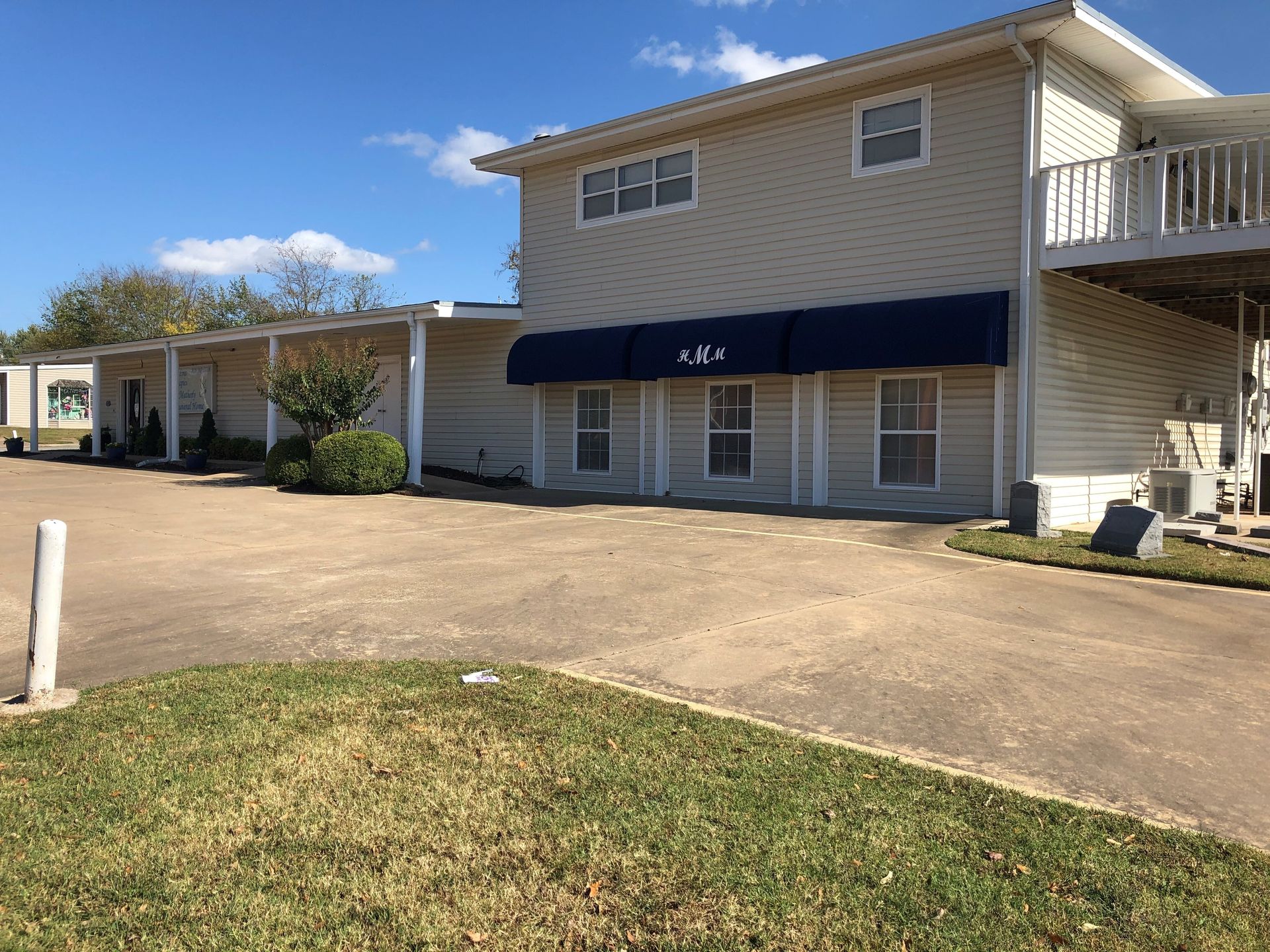 Two-story beige building with blue awning, driveway, and grassy foreground on a sunny day