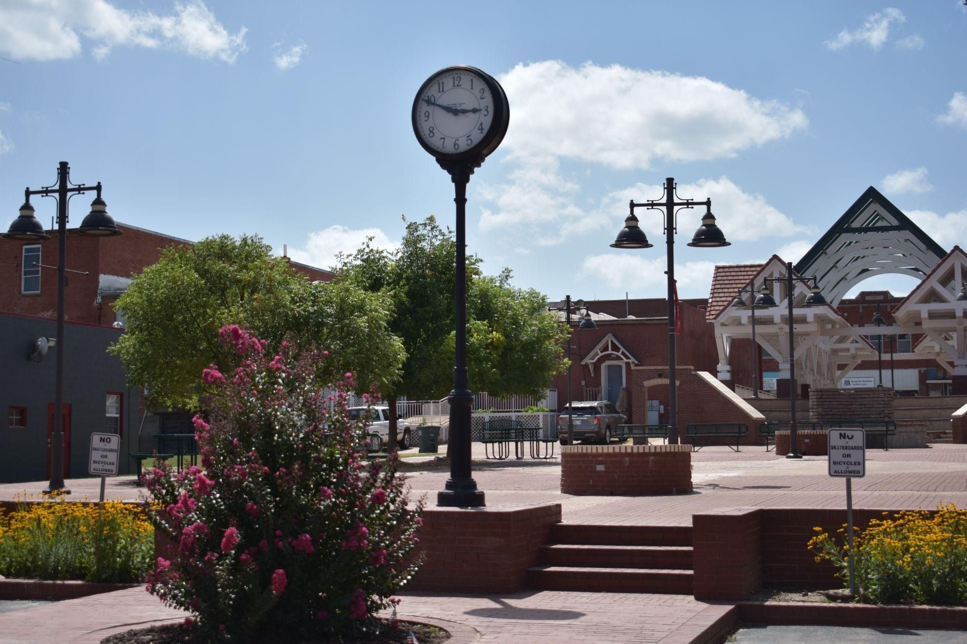 Town square with a clock, flower beds, benches, and brick buildings under a partly cloudy sky
