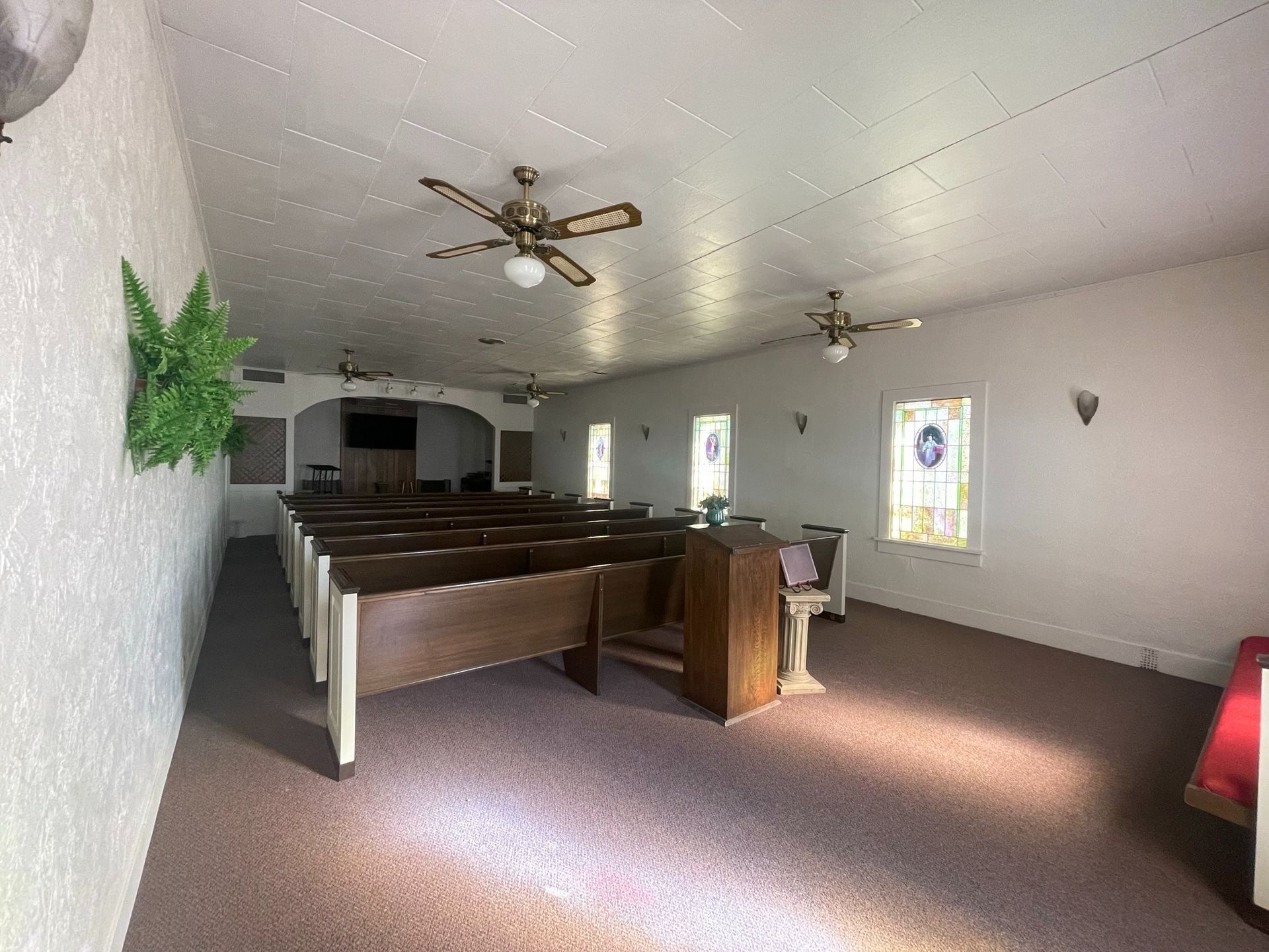 Interior of a small church with wooden pews, ceiling fans, and stained-glass windows