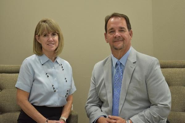 Two people seated indoors, one in a light blue shirt and one in a light gray suit, smiling at the camera.