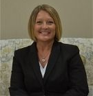 Smiling woman in a black blazer seated on a patterned couch against a beige wall