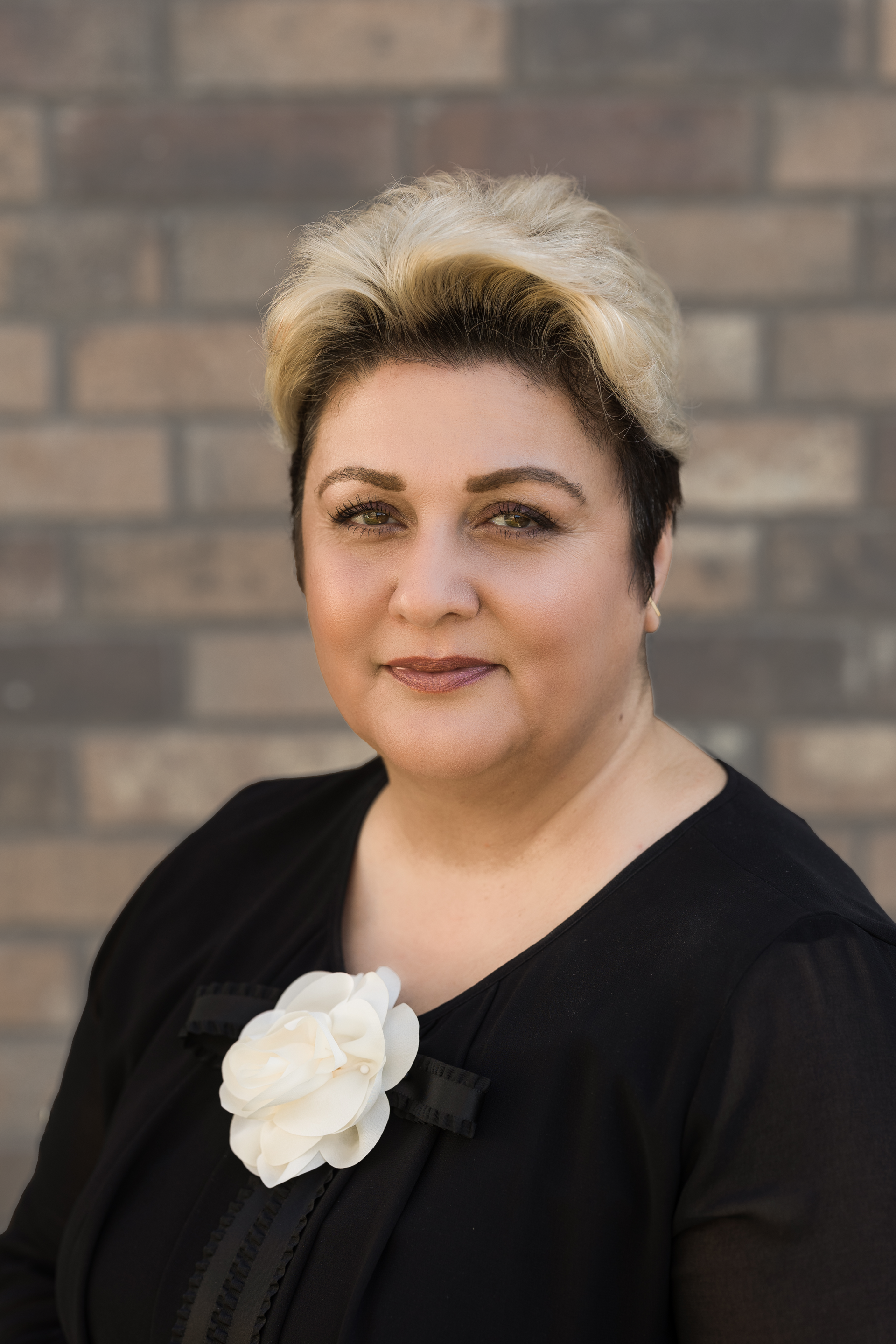 a woman in a black dress with a white flower on her collar is standing in front of a brick wall .