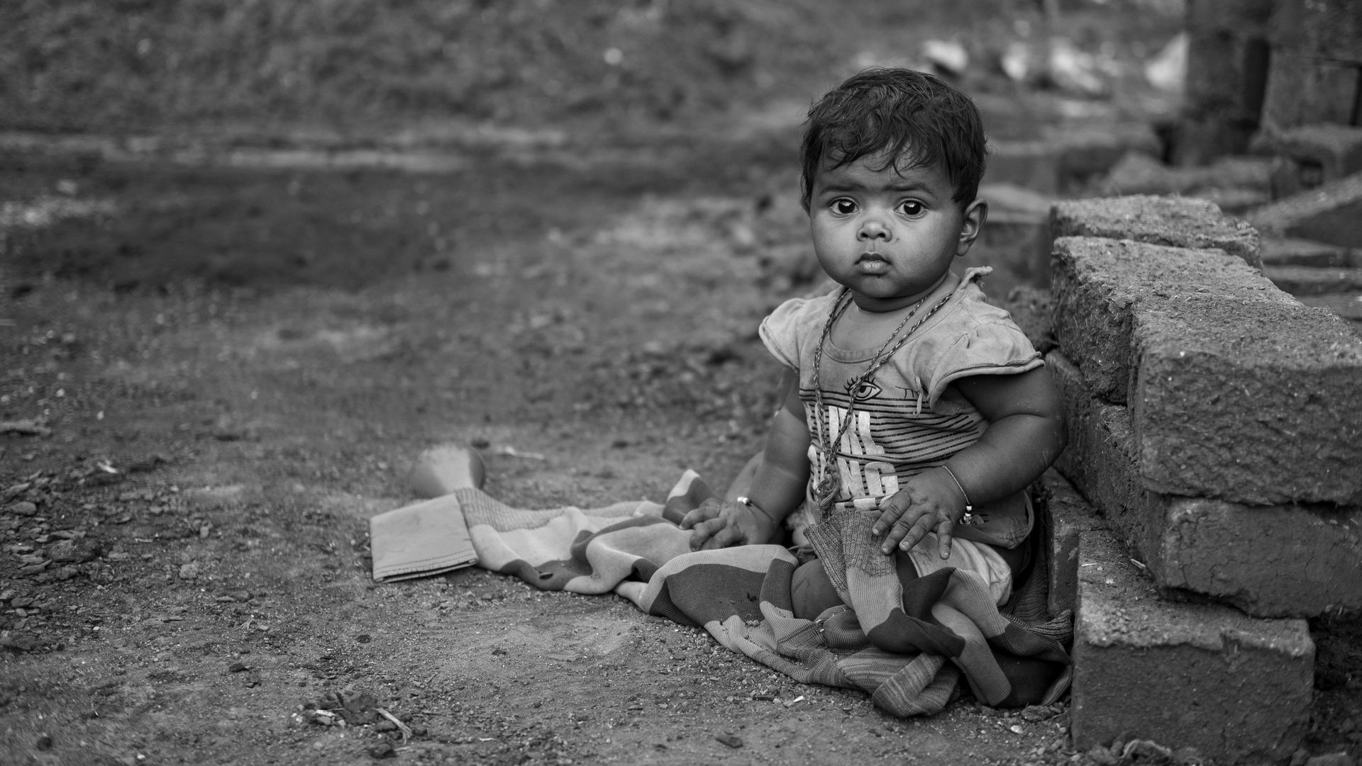A little girl is sitting on the ground next to a brick wall.