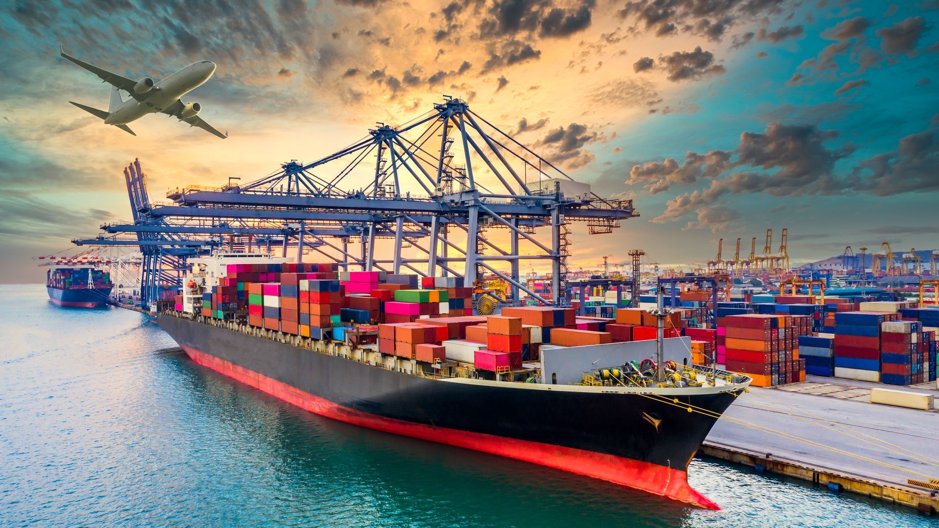 A large cargo ship is docked in a harbor with a plane flying overhead.