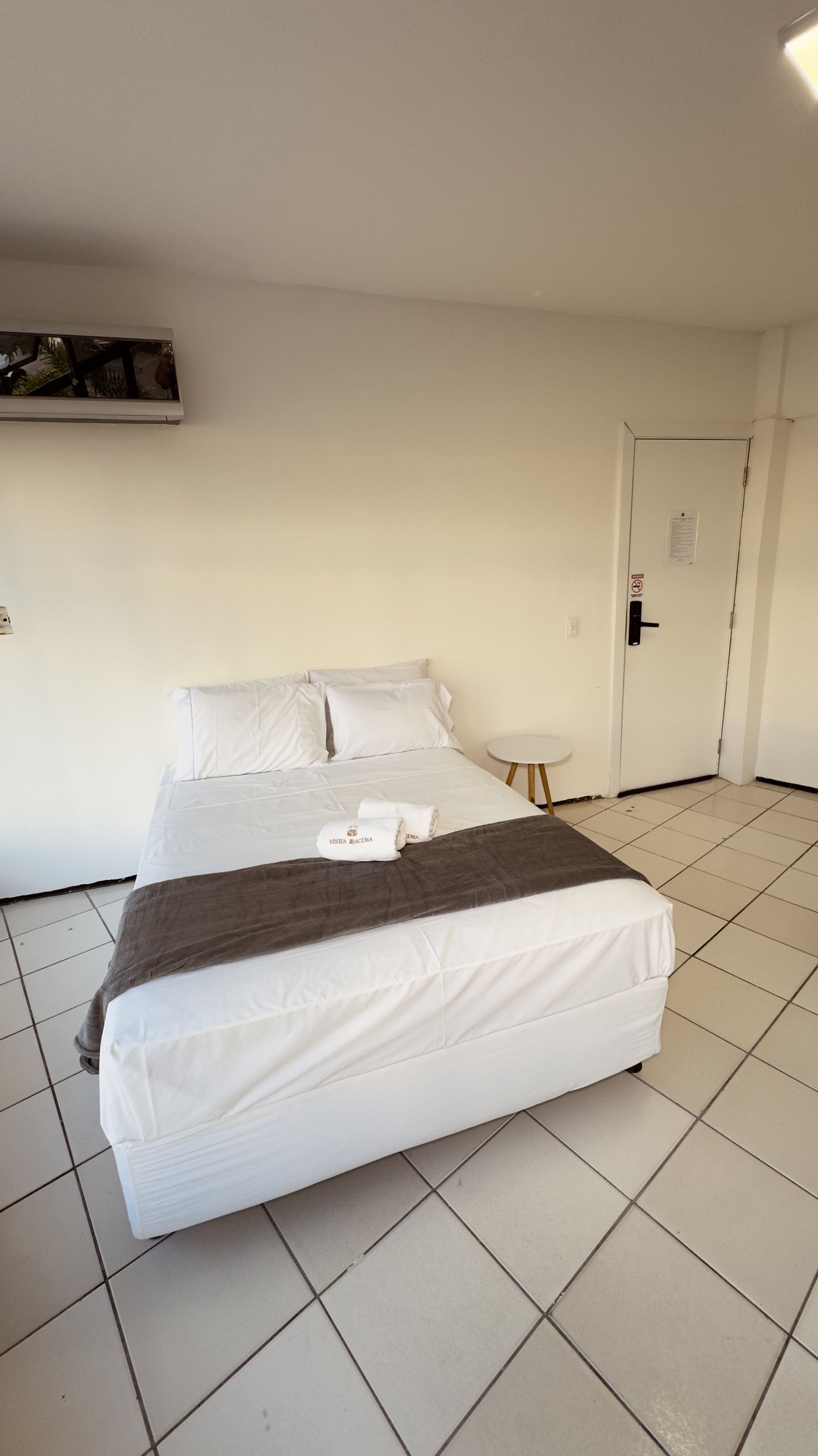 Quarto minimalista com cama branca, cobertor cinza, piso de azulejo e portas de armário fechadas.