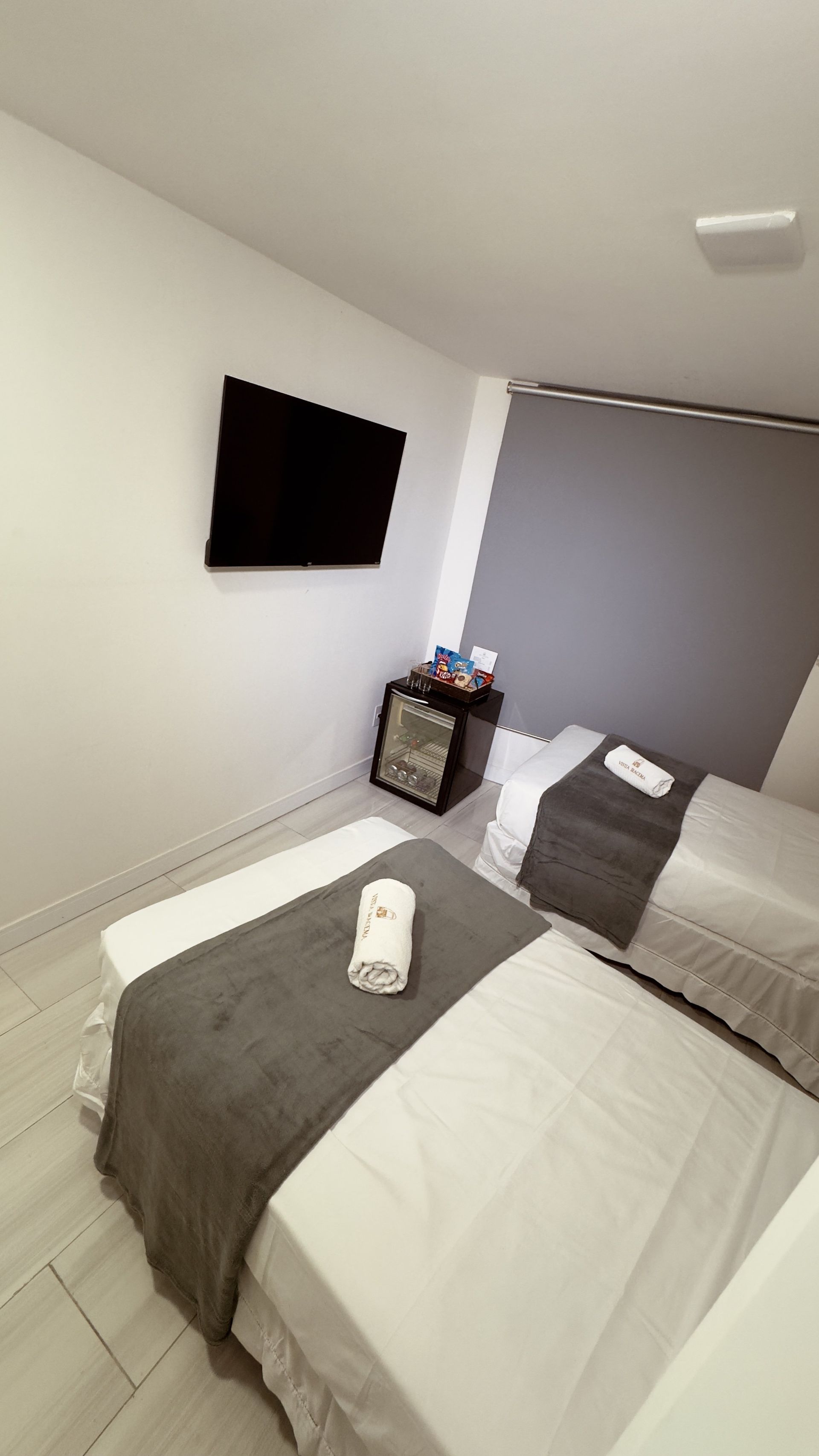 Quarto pequeno com duas camas, TV de parede, persiana e mesa de cabeceira.