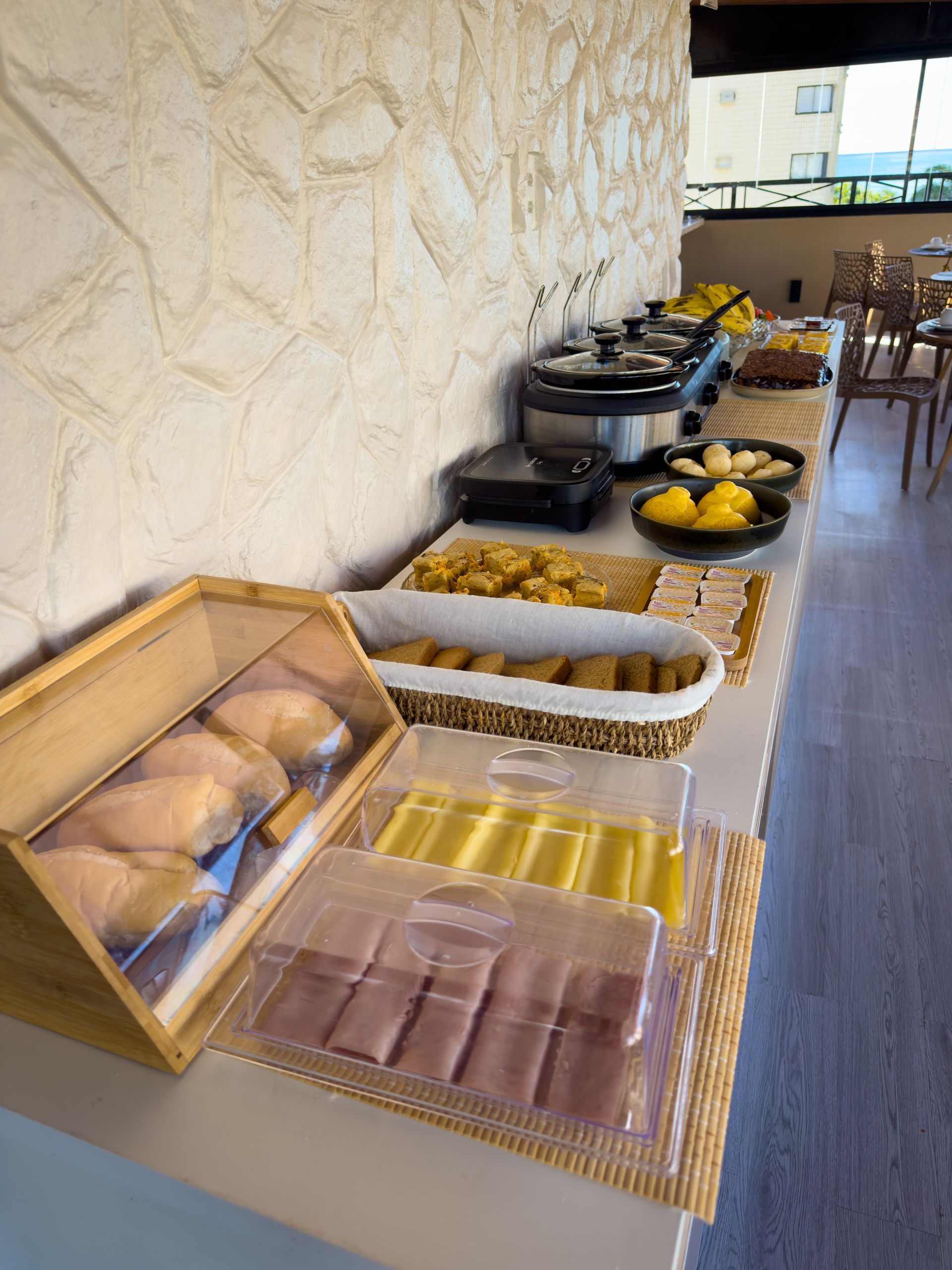Buffet de café da manhã com doces, cereais e frutas ao longo de uma parede de pedra em uma área de refeições.