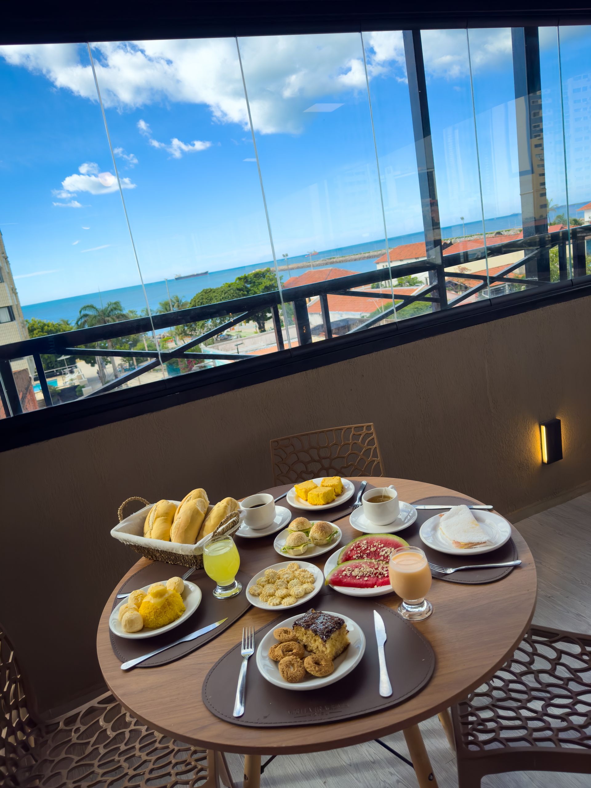 Mesa de café da manhã na varanda com doces, frutas, café e vista para o mar sob um céu azul.