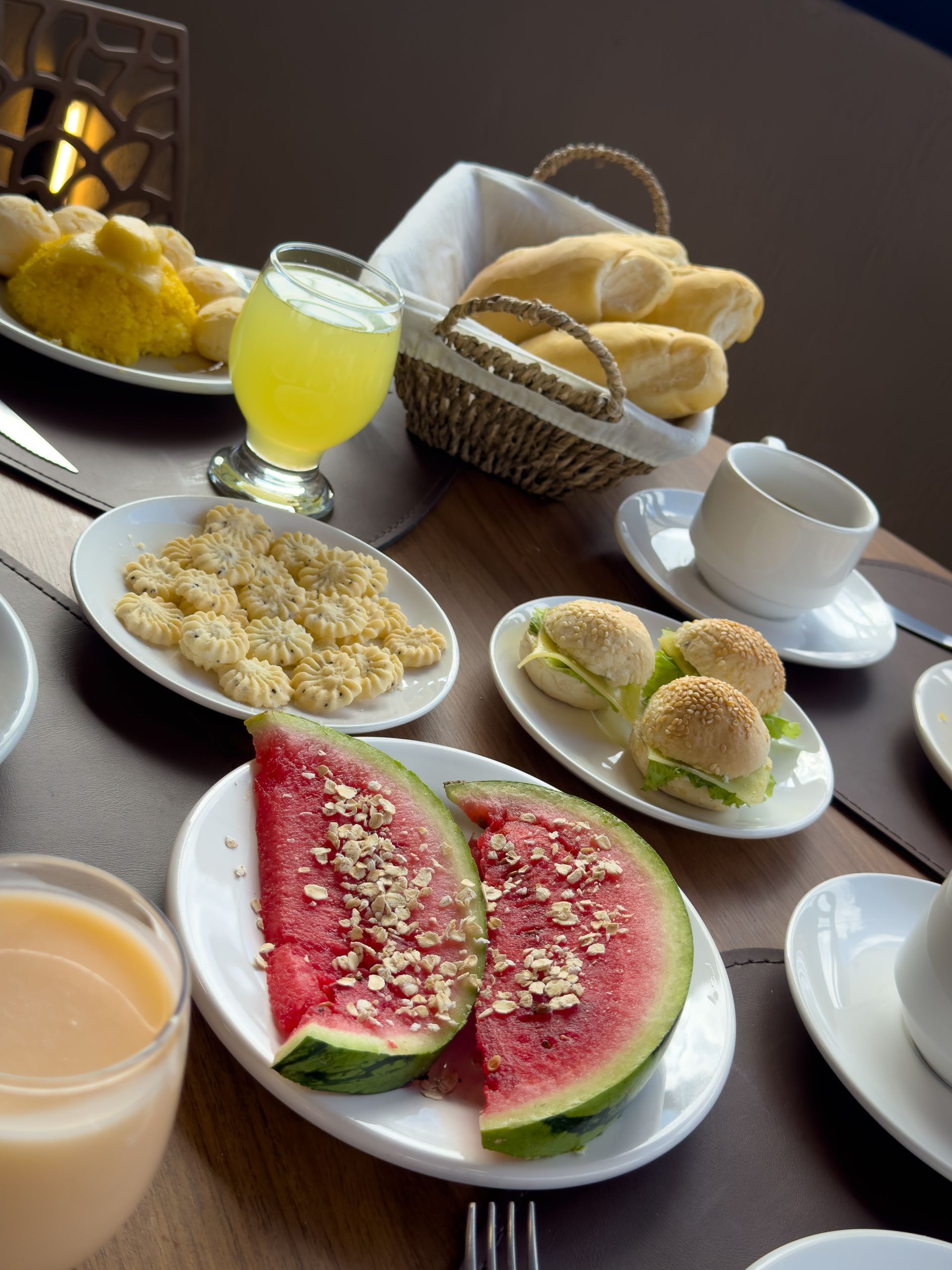 Mesa com um buffet de café da manhã contendo melancia, doces, pãezinhos, suco e café.