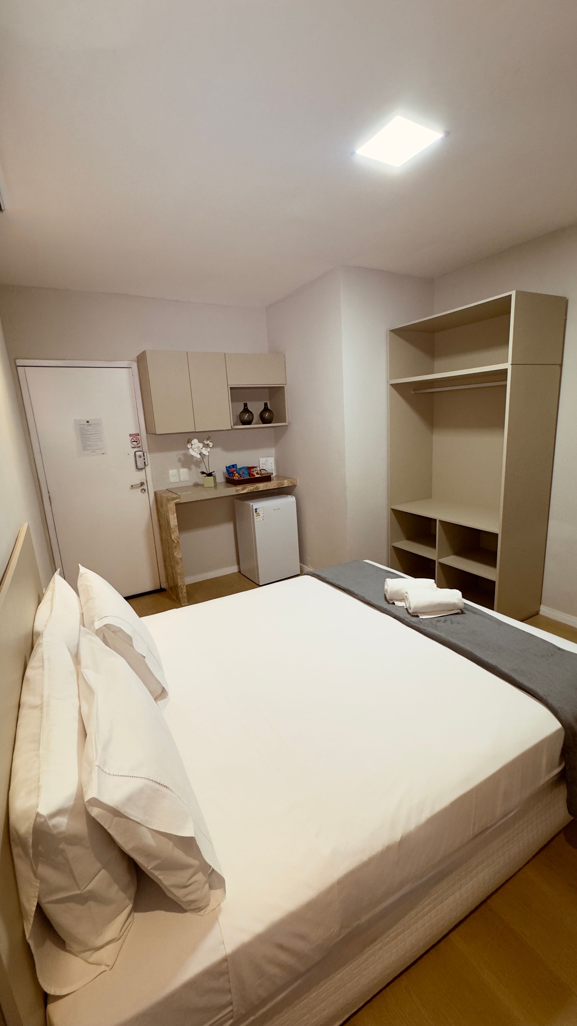 Quarto pequeno com cama, guarda-roupa aberto, escrivaninha e cozinha compacta em um apartamento moderno e iluminado.