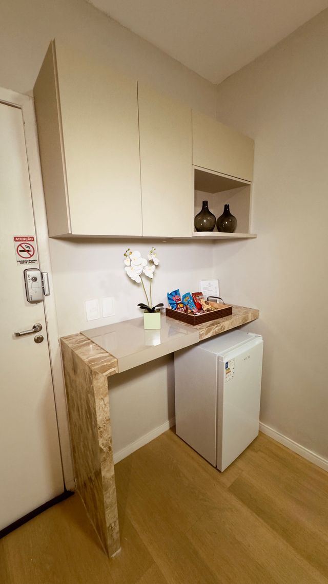 Pequeno canto de cozinha com armários brancos, um frigobar e uma escrivaninha de madeira encostada em uma parede creme.