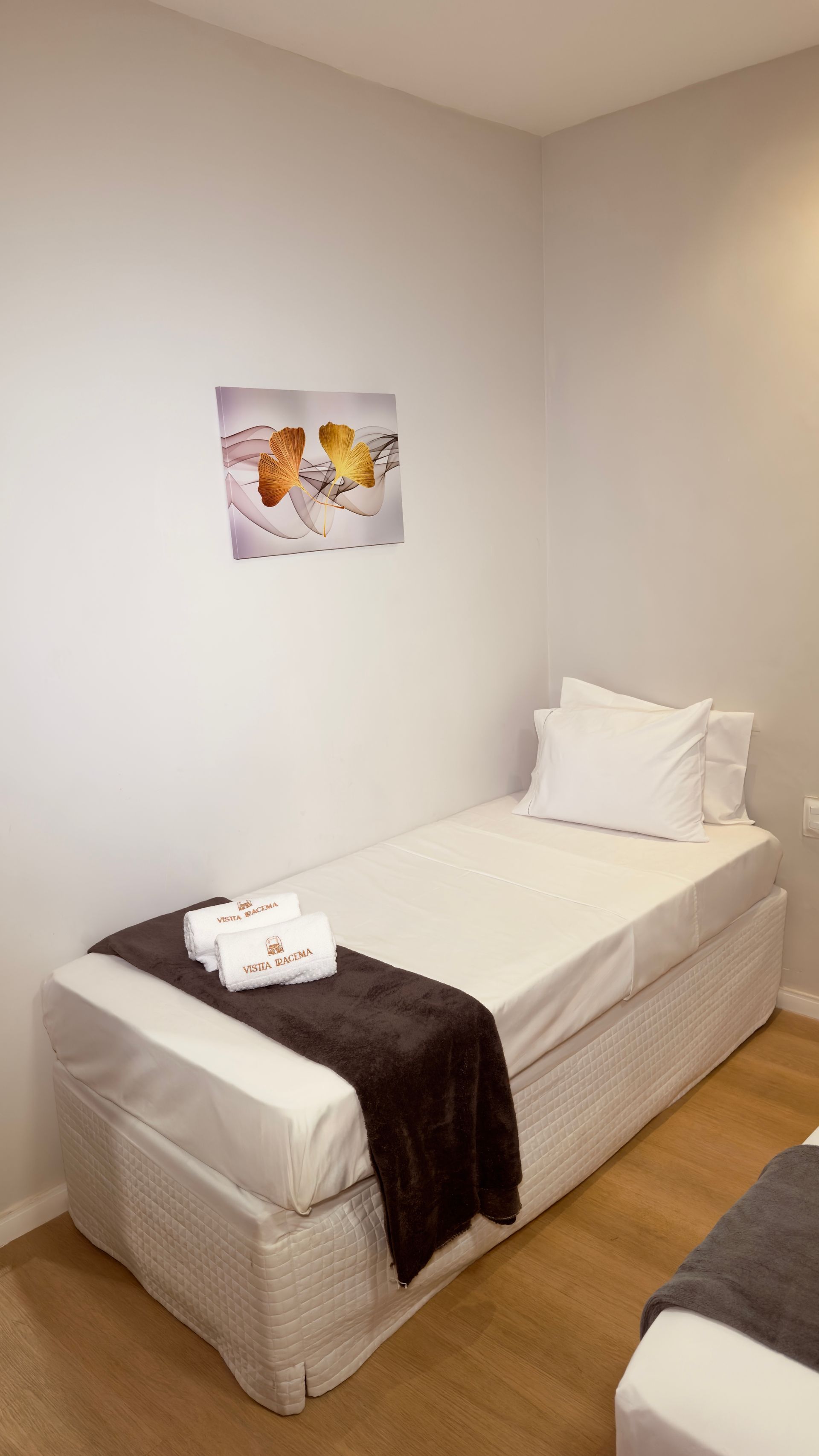 Quarto pequeno e minimalista com cama de solteiro, roupa de cama branca, manta escura e quadro de borboletas na parede.