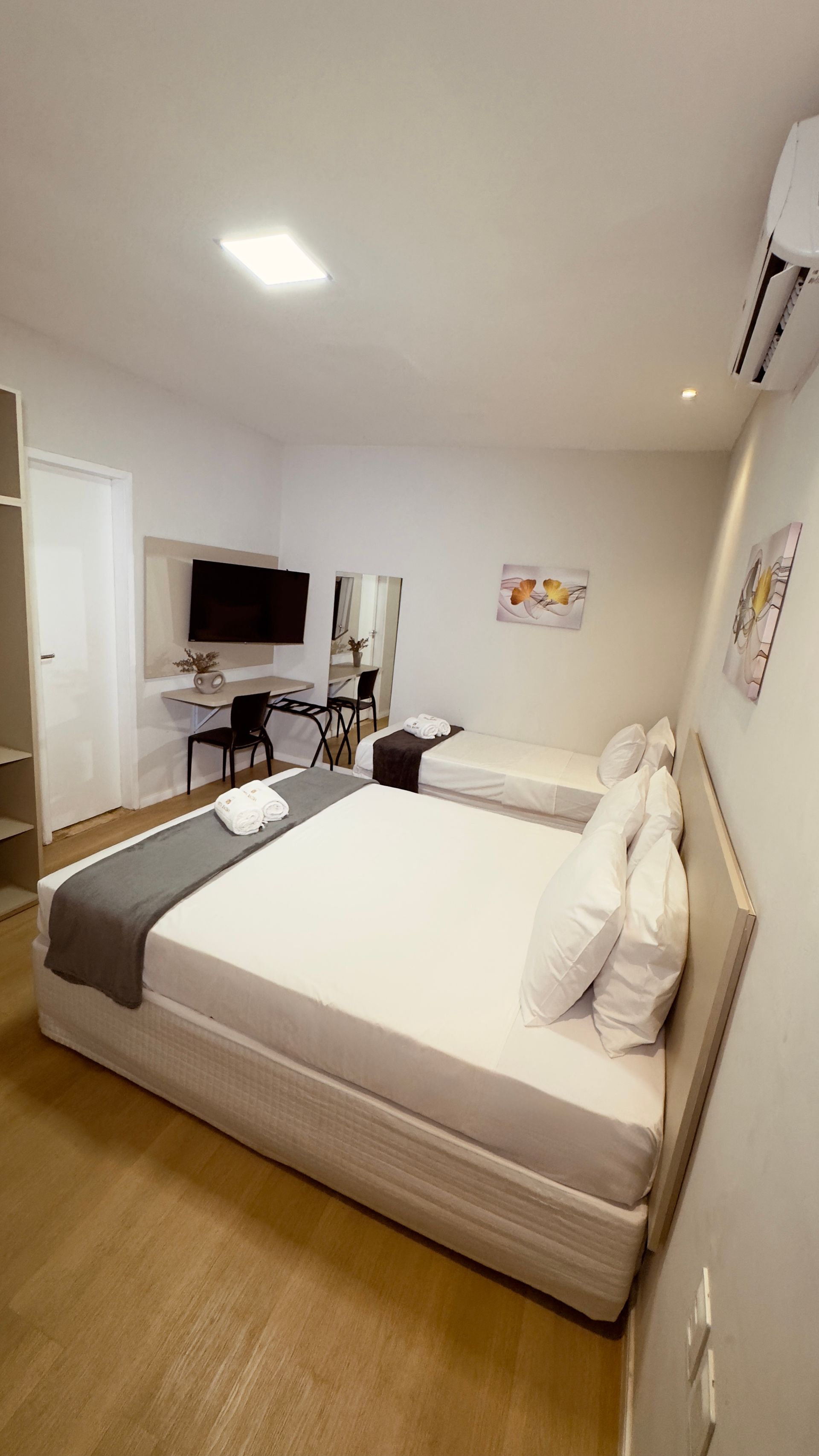 Quarto moderno com cama, TV, penteadeira e paredes brancas, visto da porta.