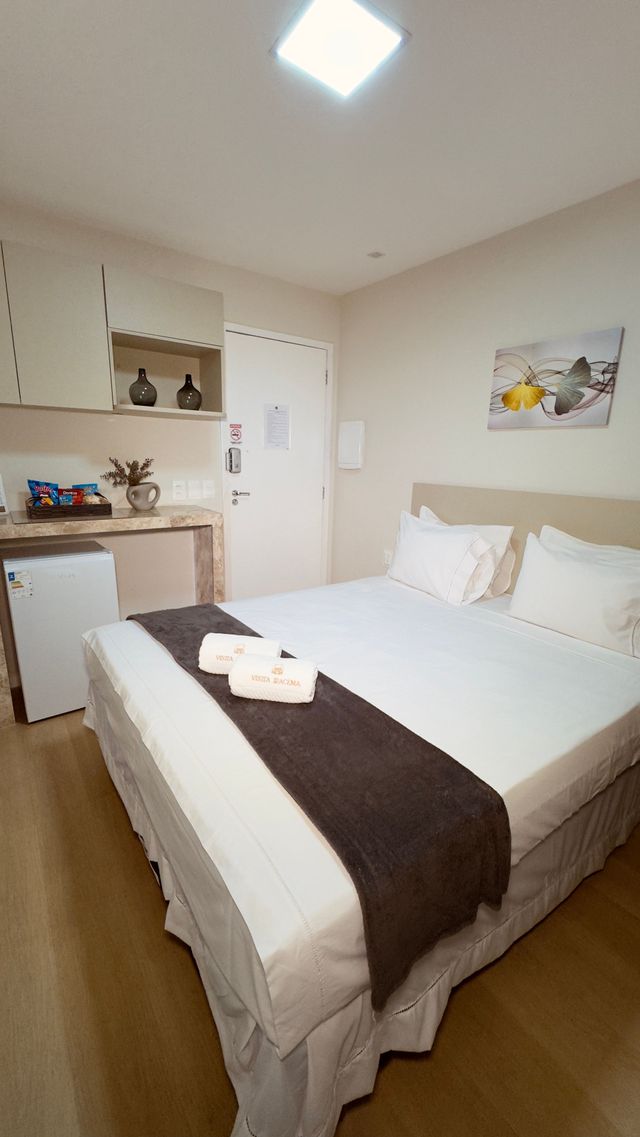 Quarto luminoso com cama arrumada, roupa de cama branca, tapete marrom e pequena cozinha no canto.