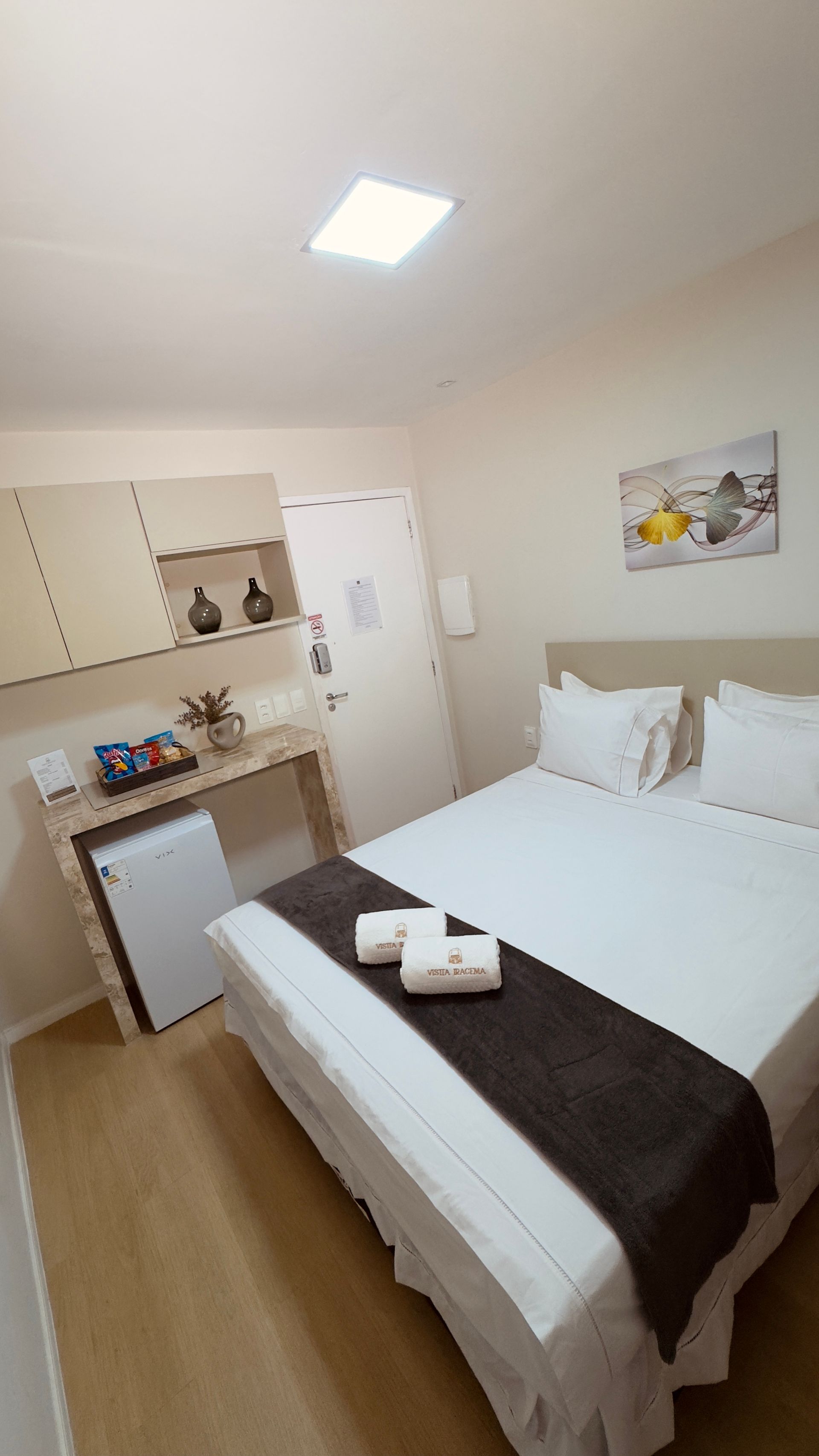 Quarto pequeno com cama arrumada, cômoda de madeira, frigobar e obras de arte na parede.