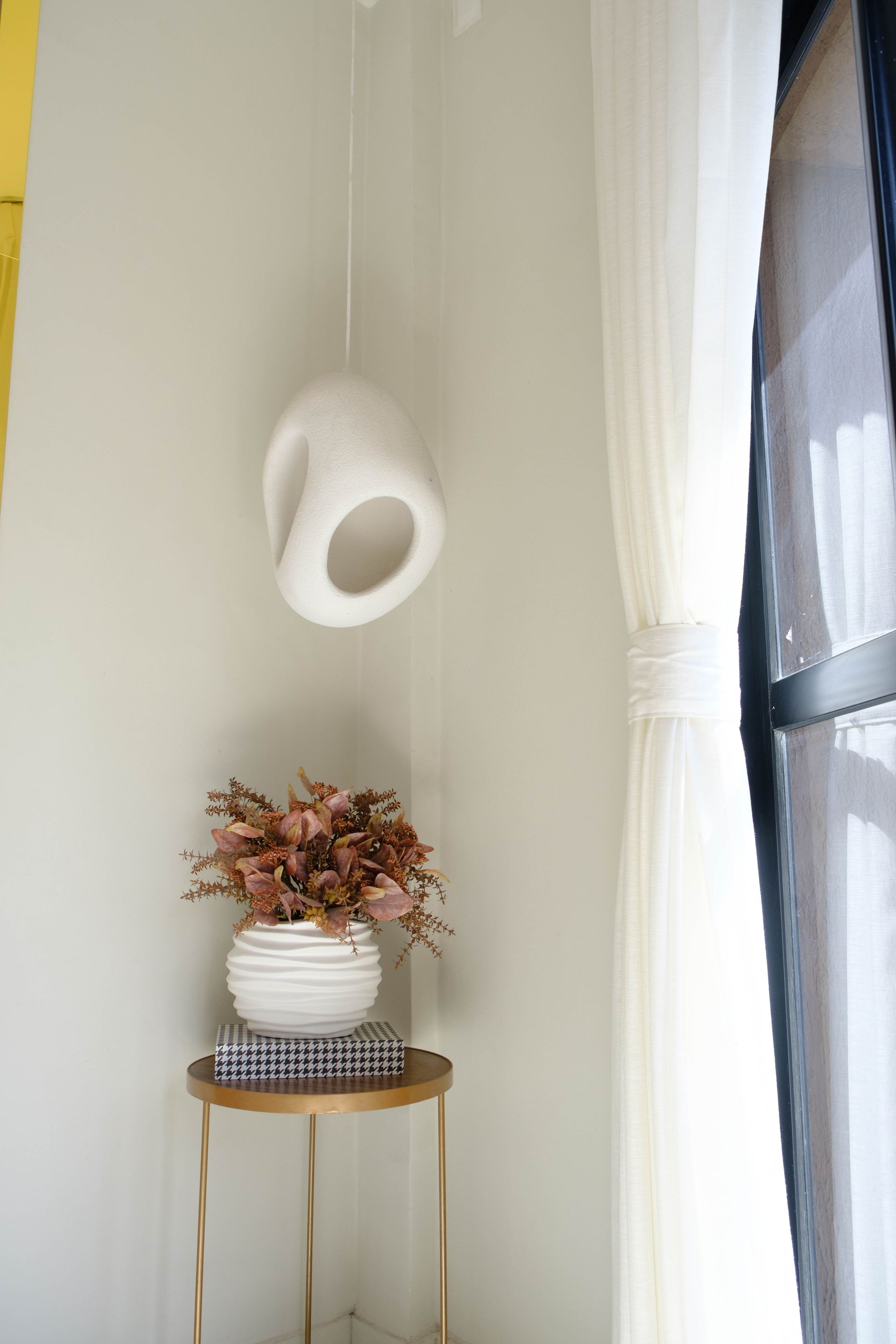 Vaso suspenso branco ao lado de uma mesa lateral dourada com um arranjo de flores secas perto da janela.