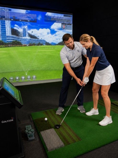 Golfer swinging a club on an indoor simulator with a large scenic screen behind him.